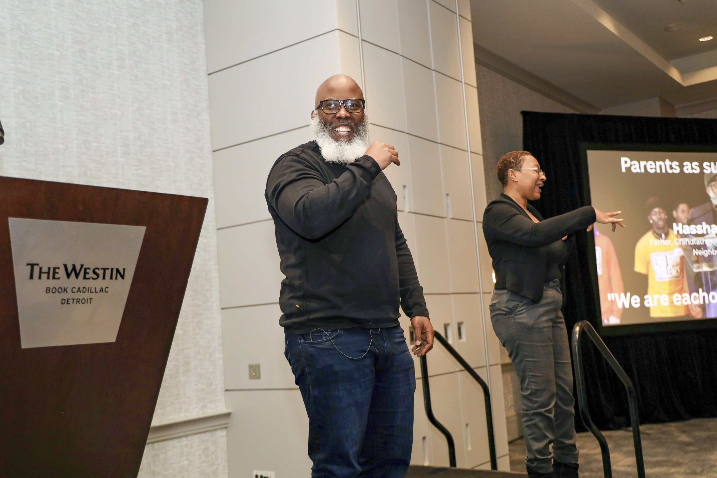 A man with glasses and a beard wearing a black shirt and jeans standing on a stage, smiling at the camera. A woman next to him is pointing towards a large screen displaying a presentation, with a podium labeled 'The Westin Book Cadillac Detroit' near
