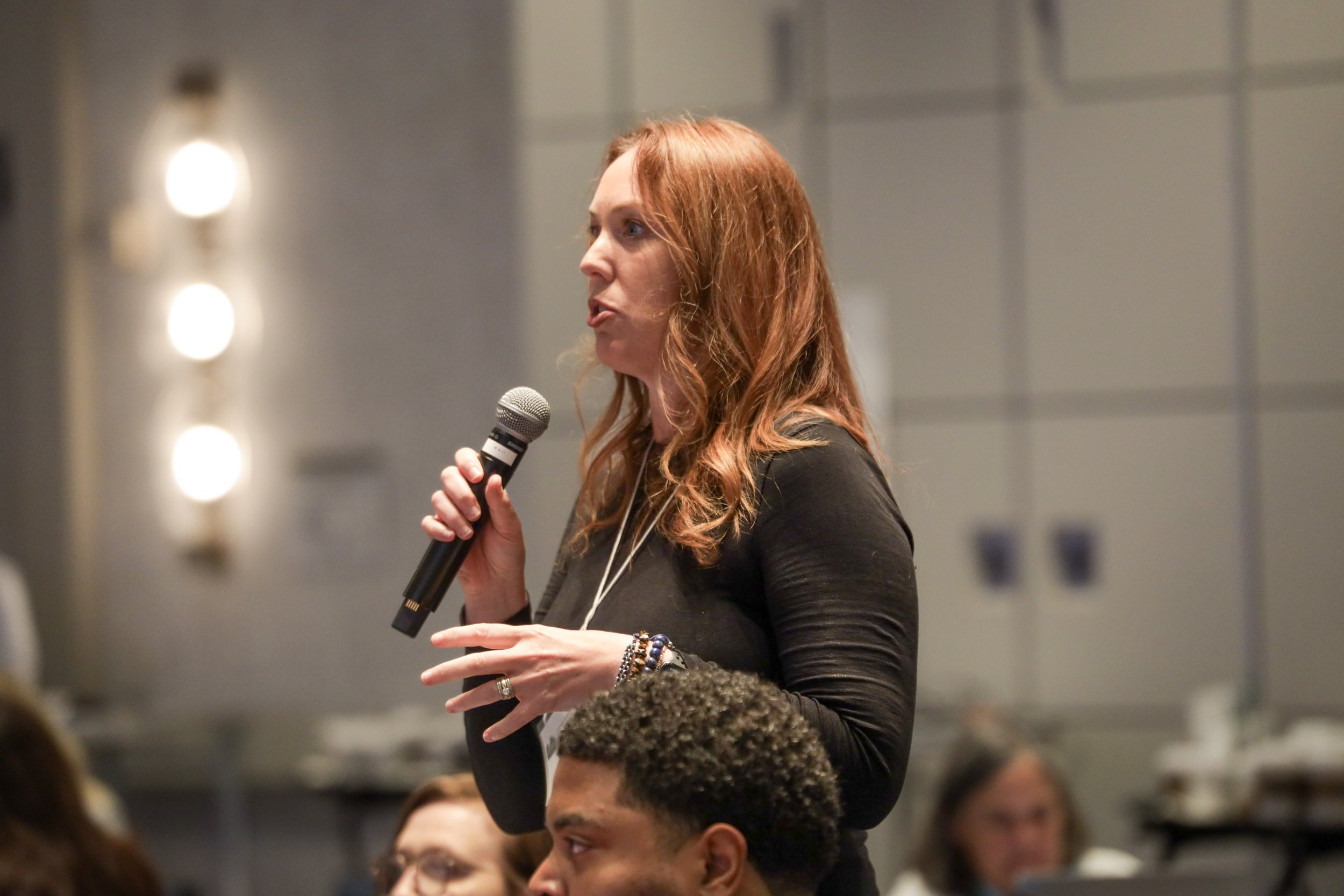 A woman with red hair speaking into a microphone at a conference or event, with people seated in front of her.