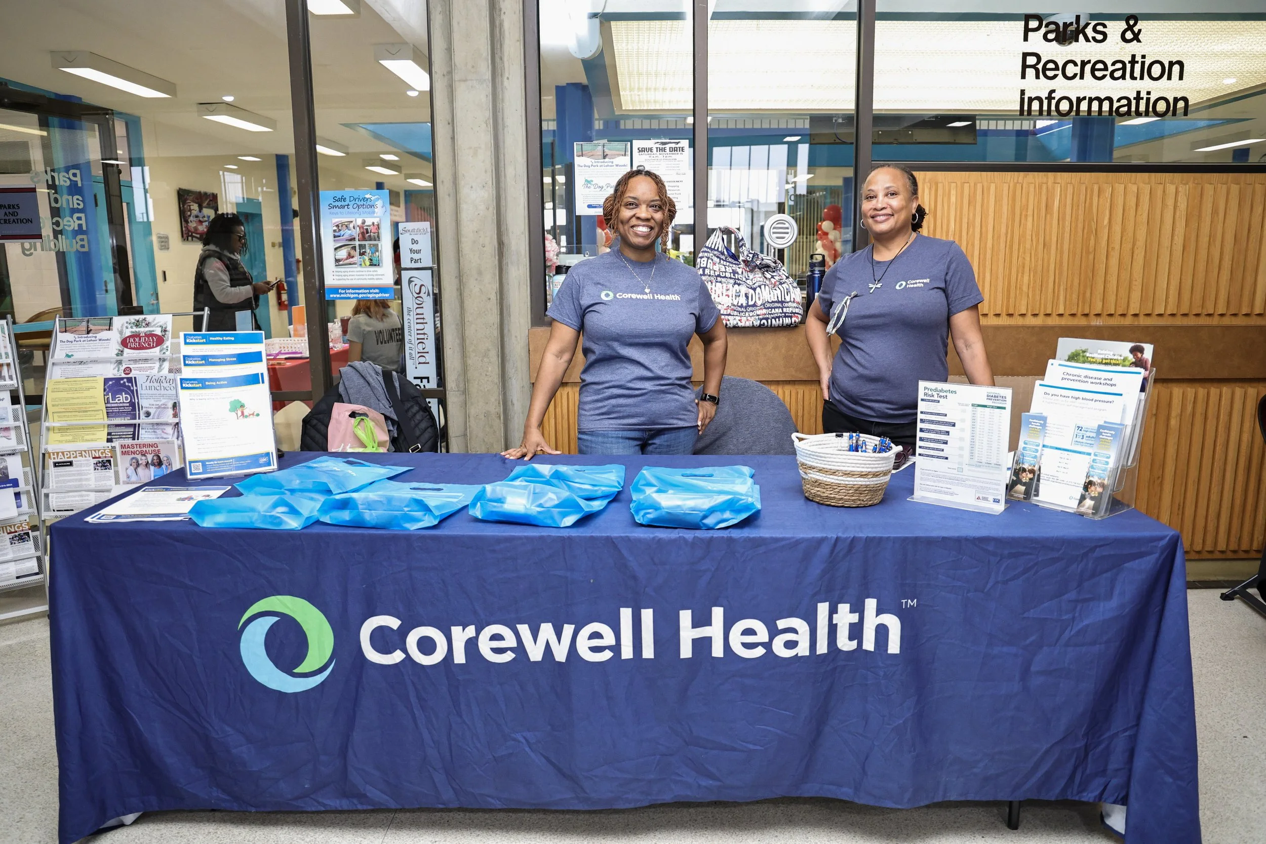 Two women standing behind a table covered with a Corewell Health cloth at a community event, with informational brochures, blue tote bags, and pens on the table in a hospital or clinic lobby.