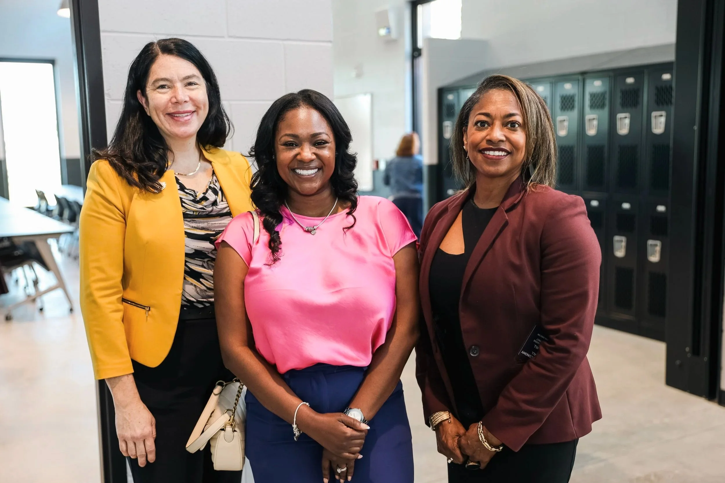 Three women standing together indoors, smiling, with lockers in the background.