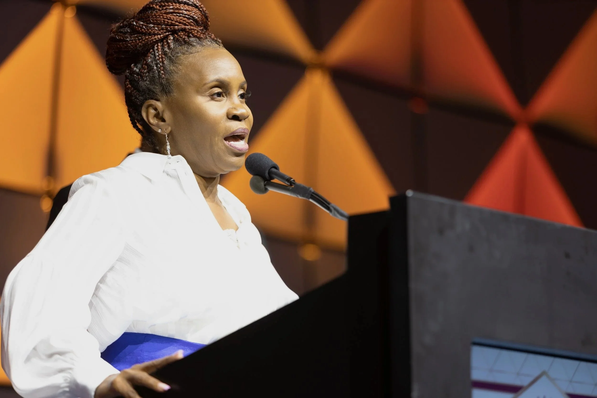 A woman with braided hair speaking at a podium with a microphone, wearing a white blouse, in front of a geometric orange and brown background.