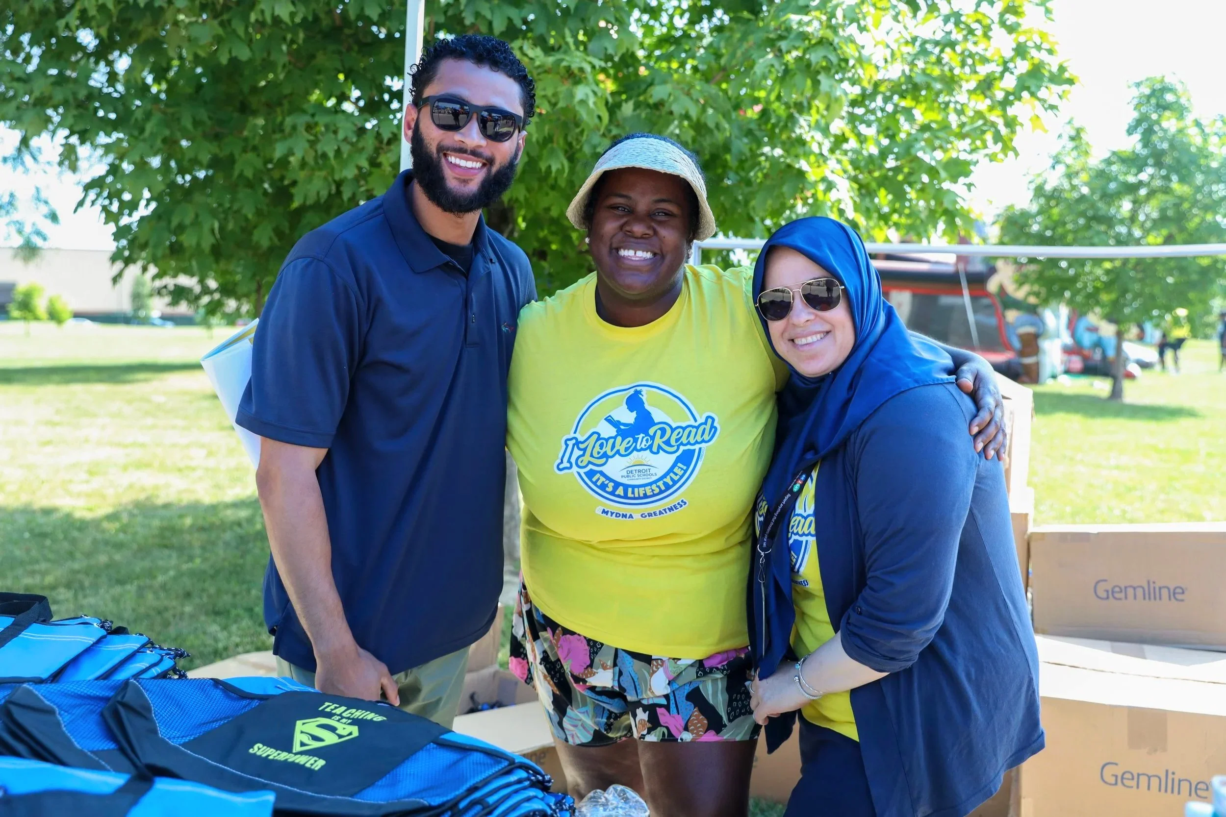 Three smiling people standing outdoors under a tree, with boxes and backpacks in the foreground, at a community event or gathering.