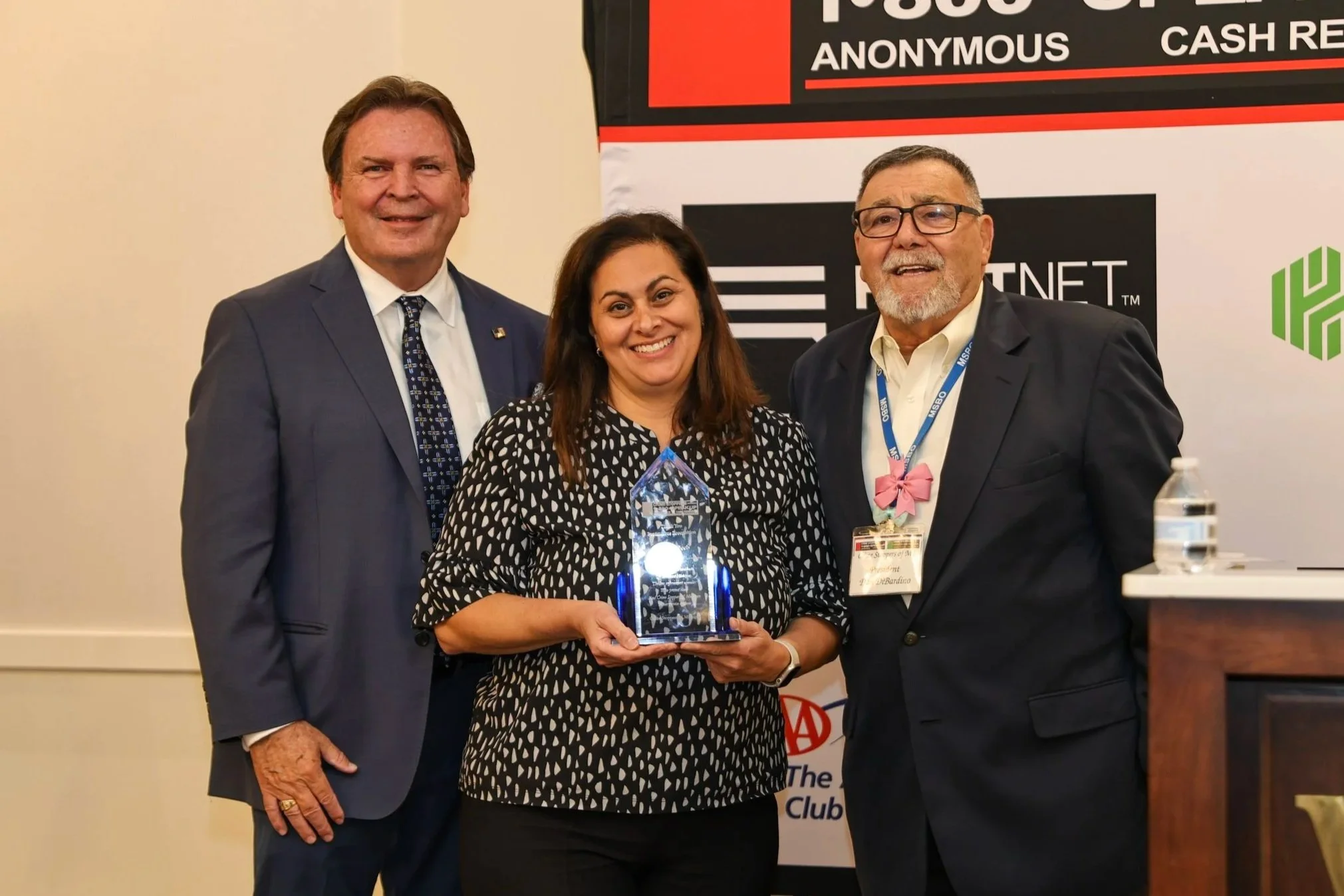 Three people standing together in formal attire at an award ceremony. The woman in the center is holding a glass award and smiling, flanked by two men. Behind them is a banner with text including "House of" and logos.