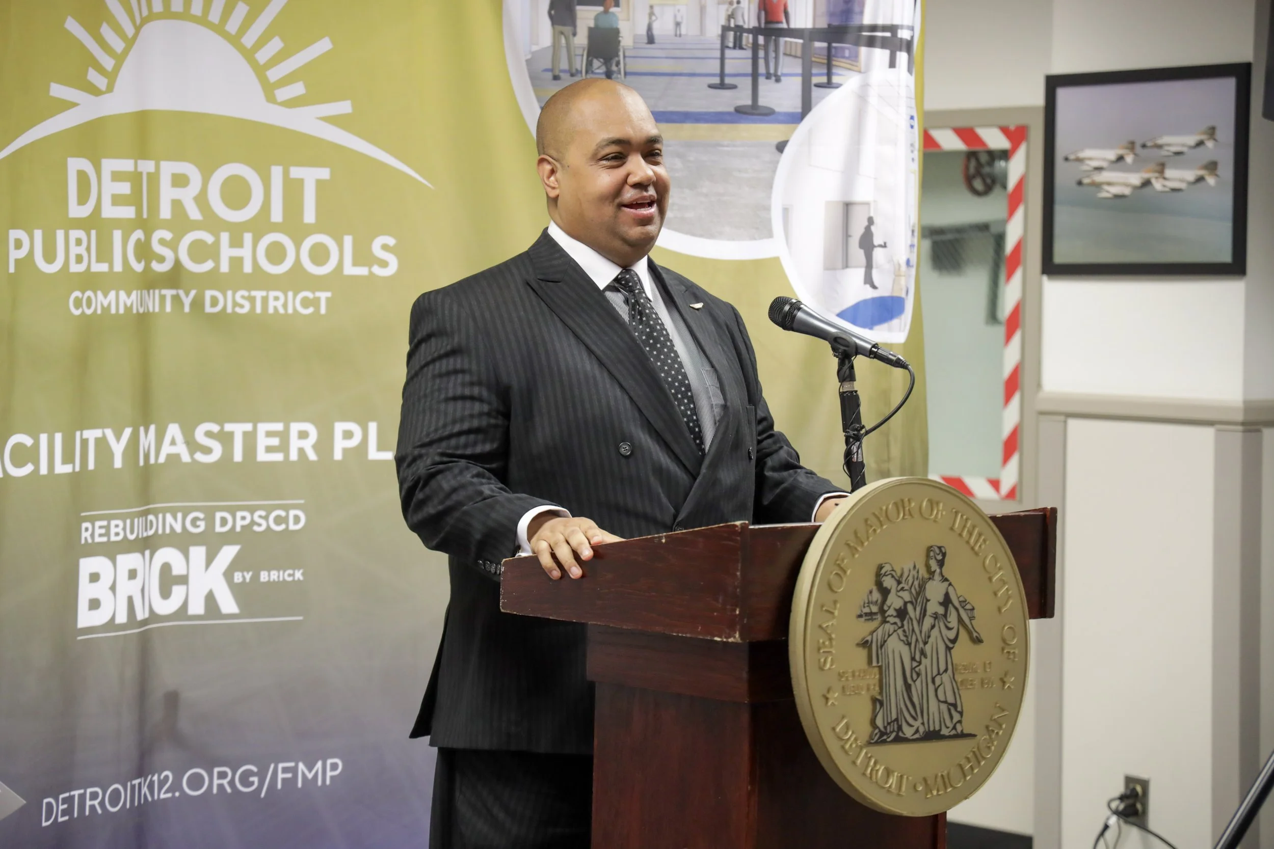 A man in a black pinstripe suit standing at a podium with the Detroit city seal, speaking into a microphone at a press conference or event