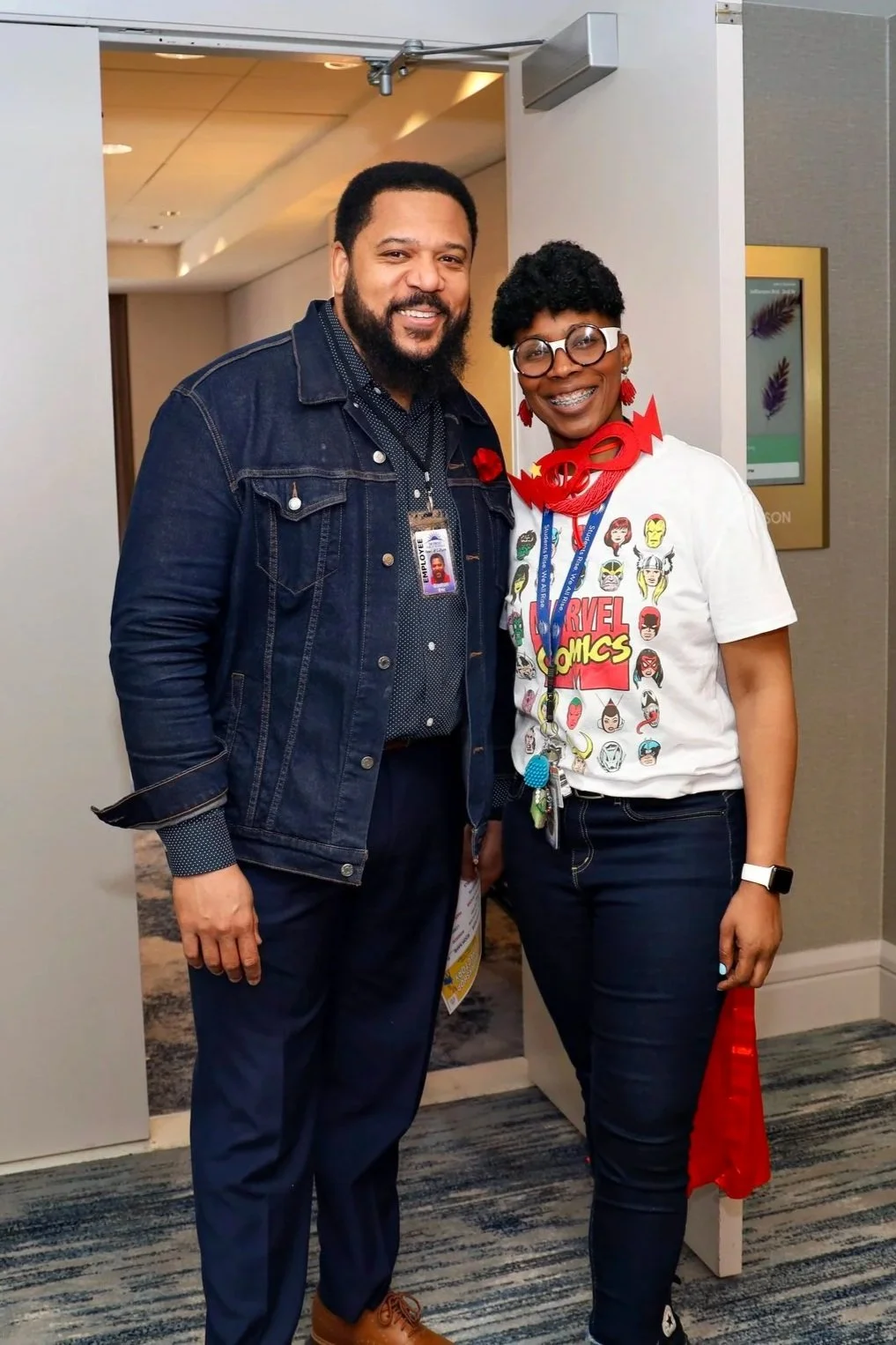 Two people standing indoors, smiling at the camera. One is a man with a beard, wearing a denim jacket, dark shirt, and blue pants. The other is a woman with short curly hair, glasses, wearing a Marvel Comics T-shirt, black pants, and a red necklace.