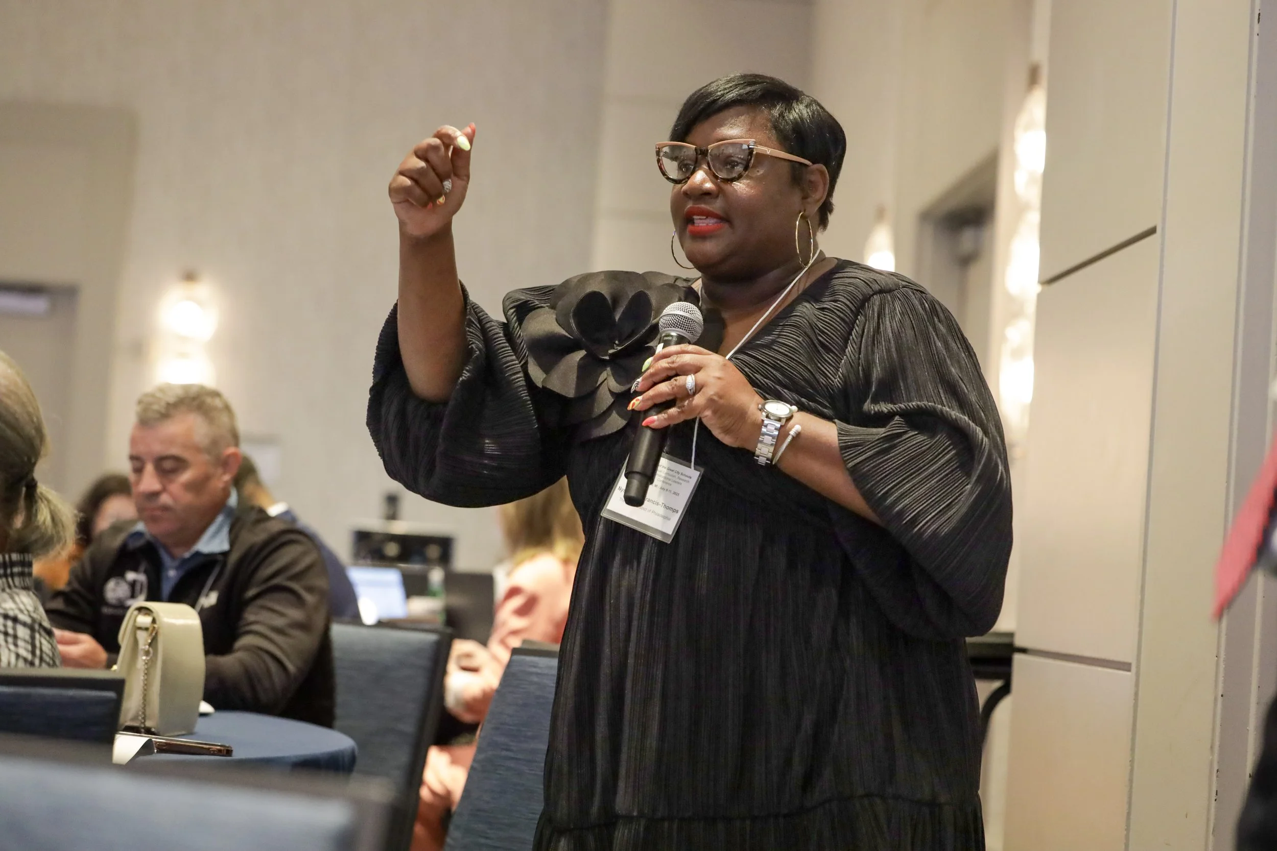 A woman with short black hair, glasses, and dressed in black, speaking into a microphone during a conference or seminar, with seated attendees in the background.