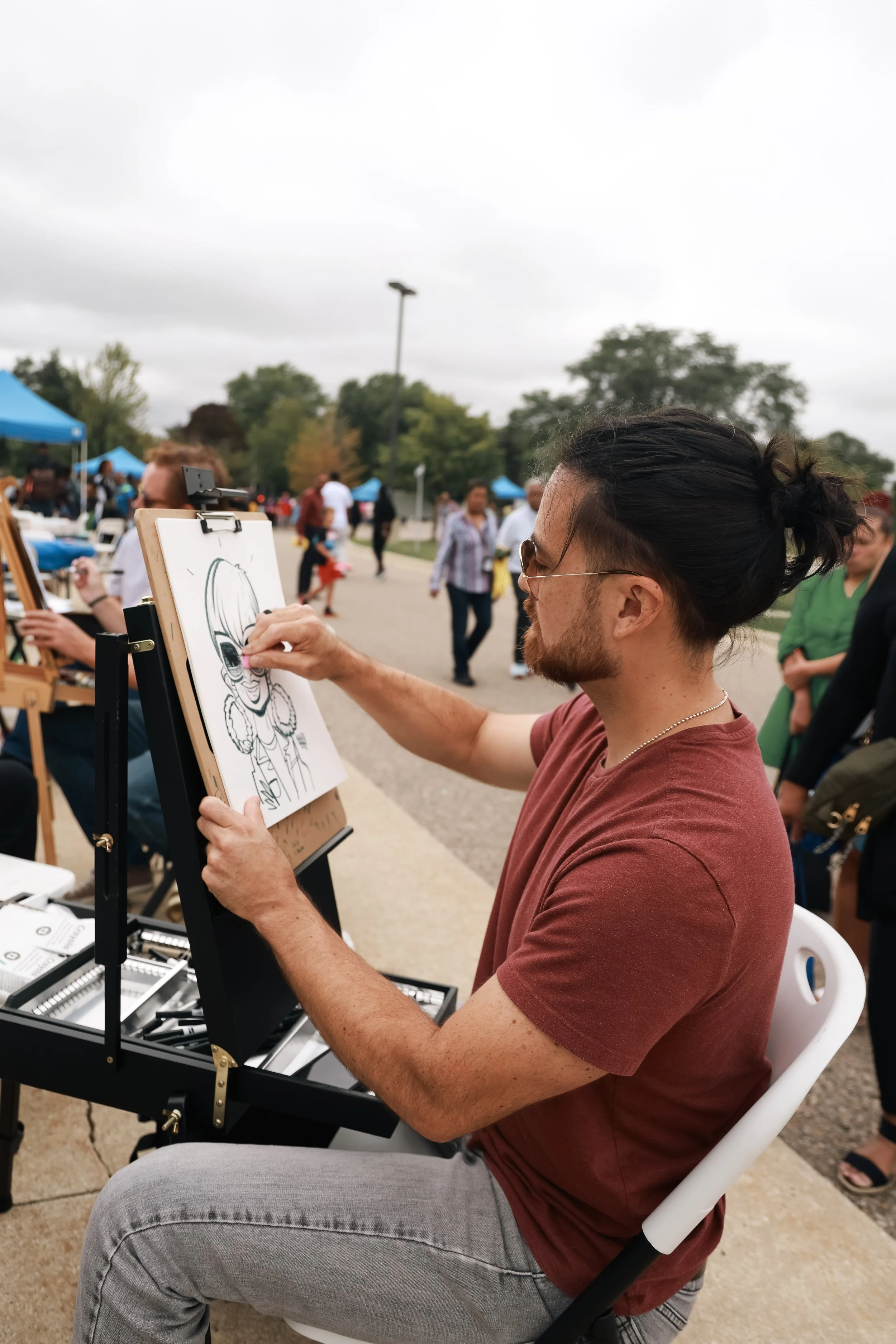 A man with glasses and a beard, wearing a red t-shirt, is drawing a caricature of a person at an outdoor event. The man has long dark hair tied back and is seated on a white chair, working on a drawing board with art supplies nearby. There are tents 