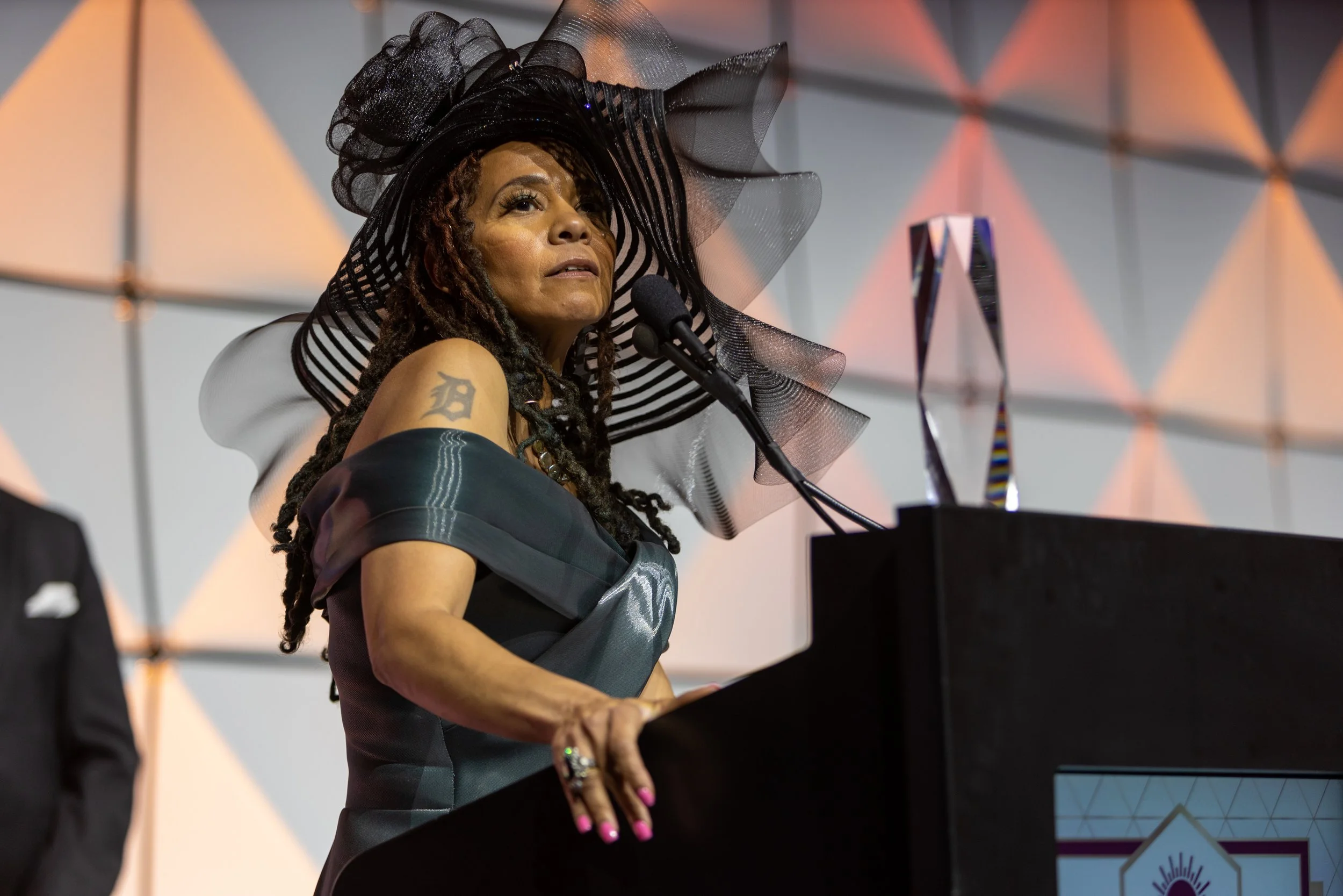 A woman with long dreadlocks wearing an elaborate black and gray dress and a large, striped black and gray hat with sheer accents speaking at a podium, with a geometric background and a trophy visible on the podium.