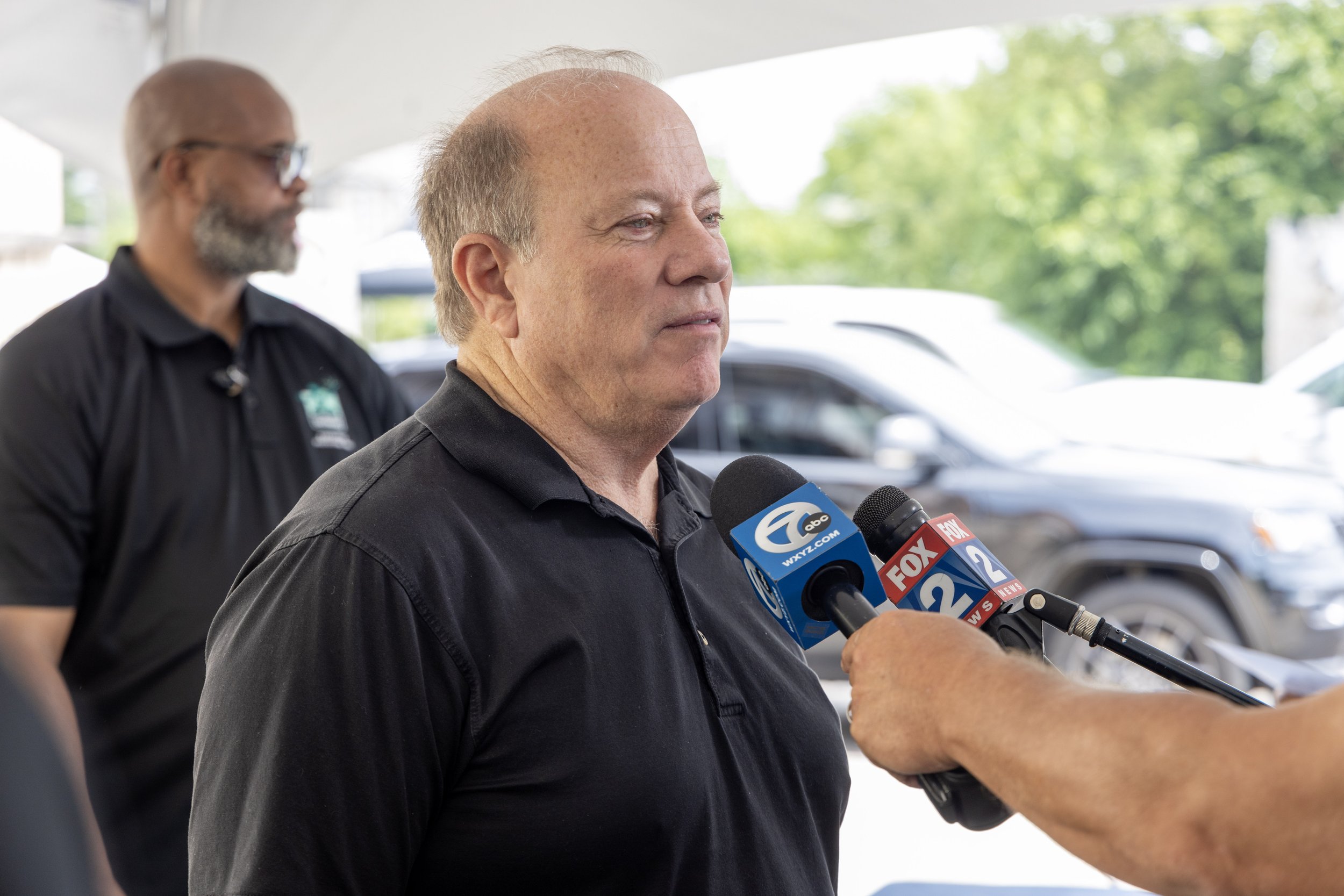 A man being interviewed by reporters outdoors, with multiple microphones in front of him, and another man standing in the background.
