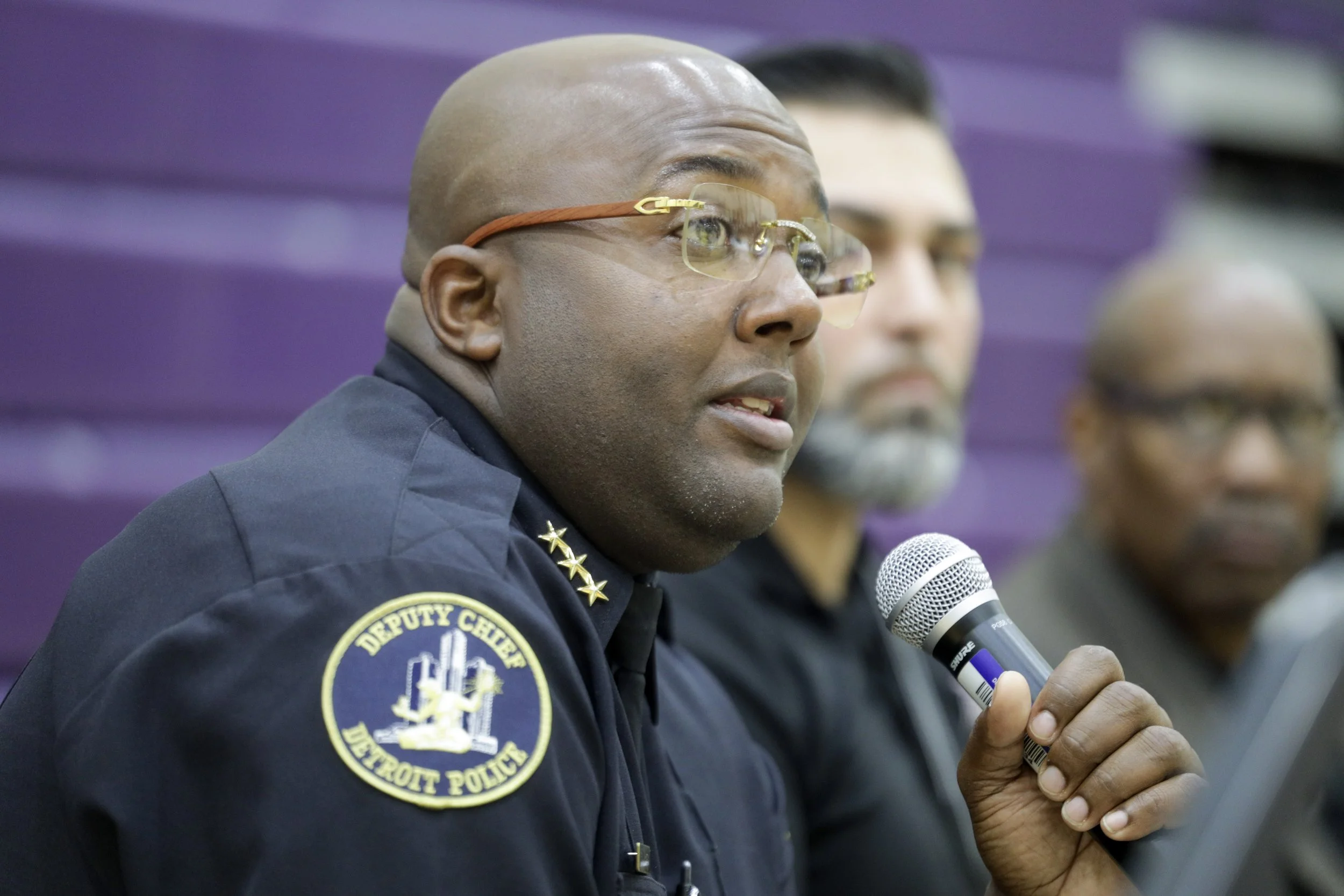 A man in a Detroit Police Department Deputy Chief uniform speaking into a microphone during a panel discussion, with two other men seated behind him against a purple background.