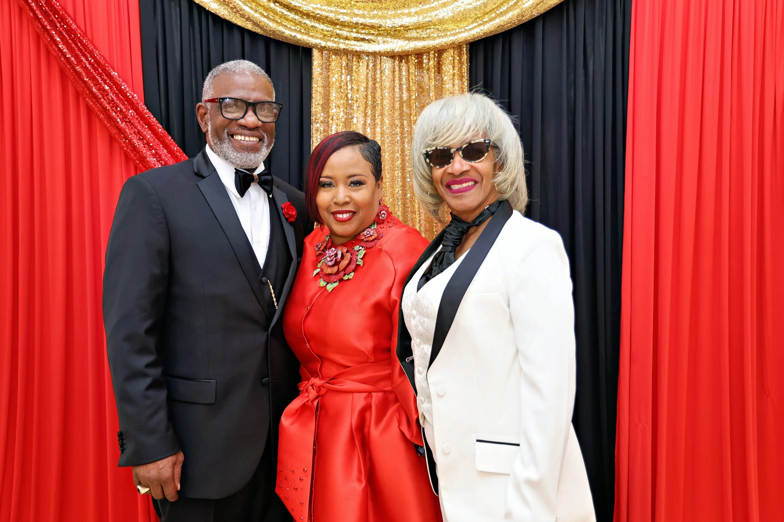 Three well-dressed older adults smiling at a formal event, standing in front of a red and black curtain with gold accents.