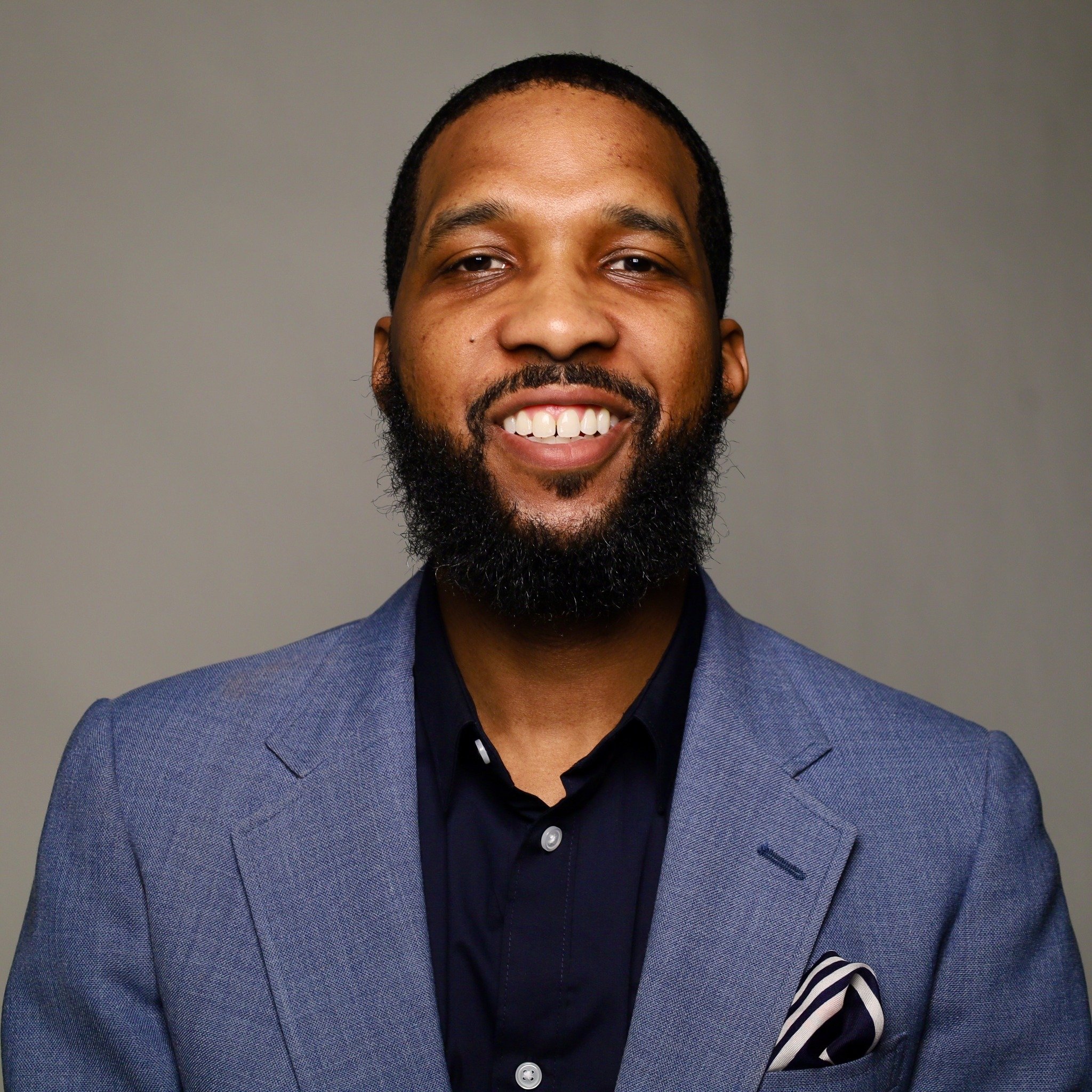 Portrait of a smiling man with a beard, wearing a blue suit jacket and dark shirt, against a gray background.