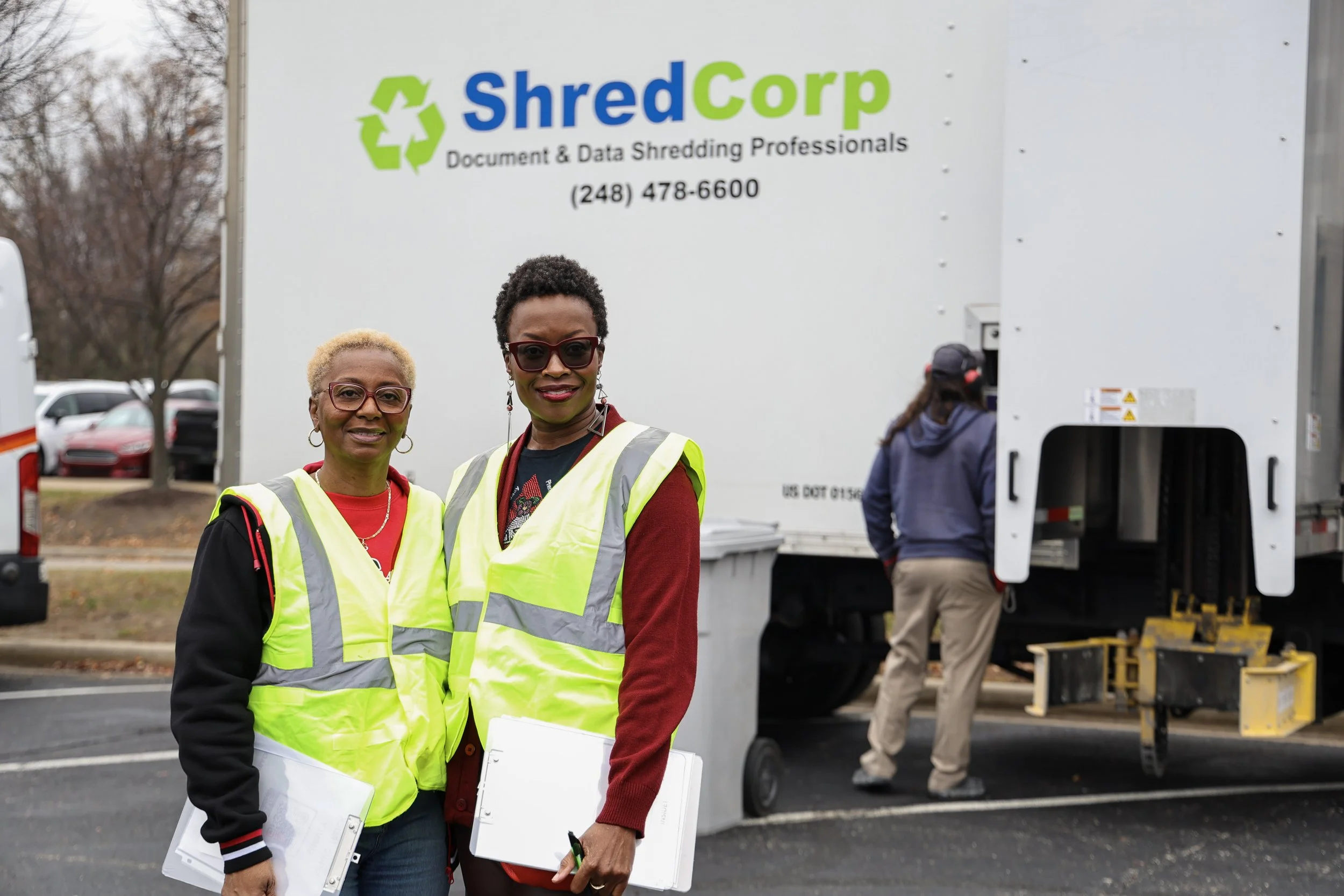 Two women wearing yellow safety vests standing in front of a shredding truck labeled ShredCorp, with another person visible in the background near the truck.