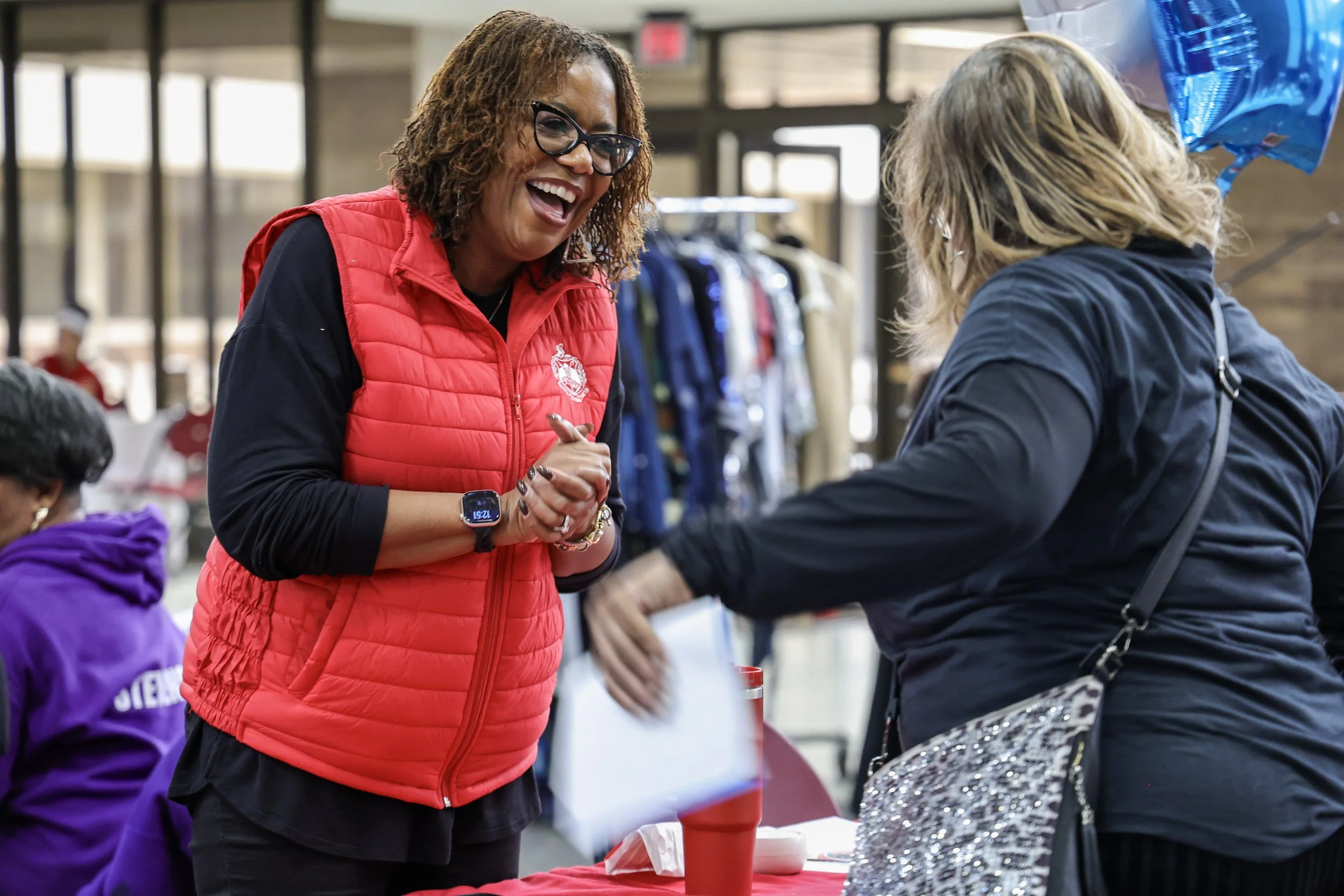 A woman in a red vest is smiling and talking to another woman at a table, in what appears to be a retail or community event setting.