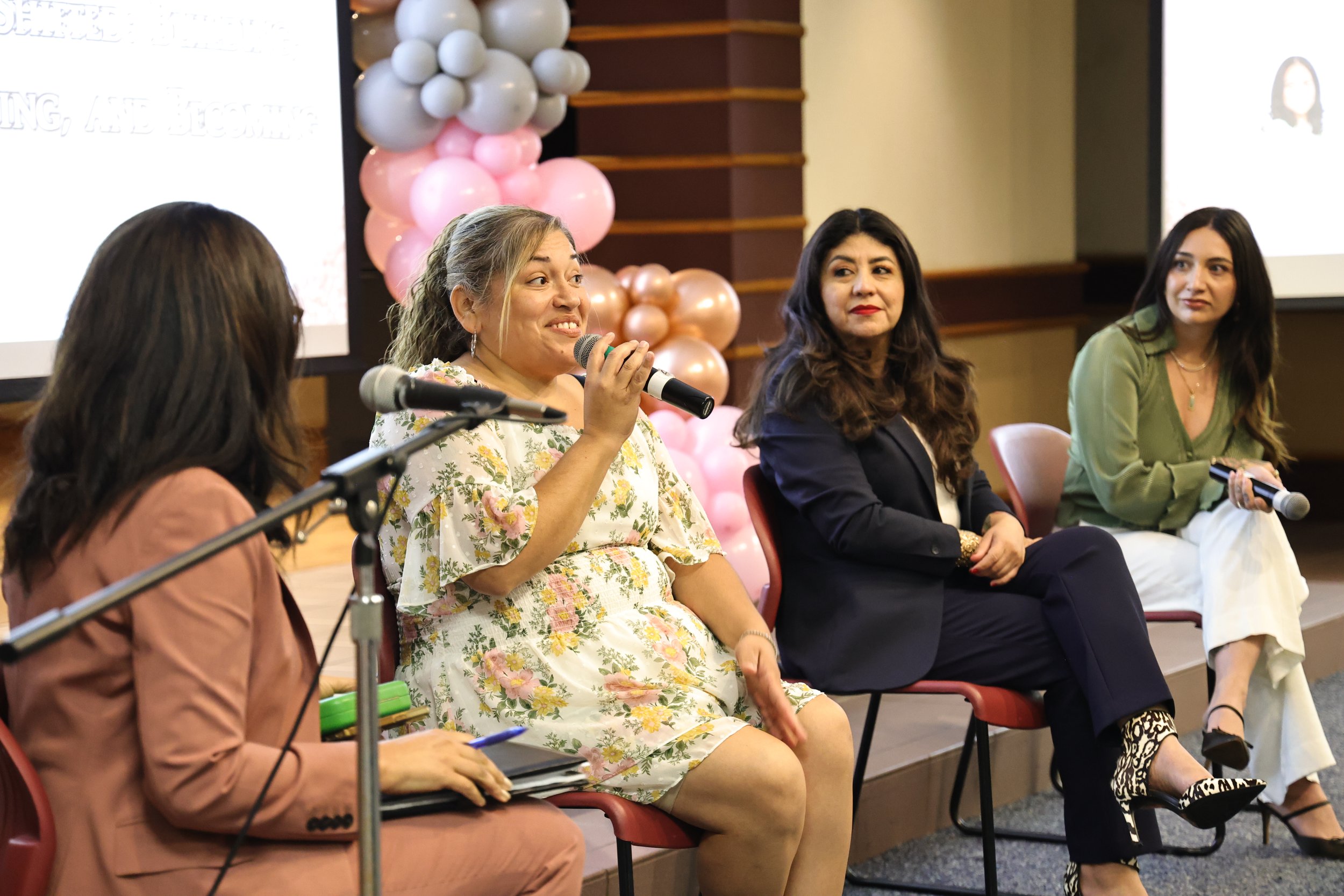 Four women sitting on chairs participating in a panel discussion. The woman second from the left is speaking into a microphone. Behind them is a pink balloon decoration and a presentation screen.