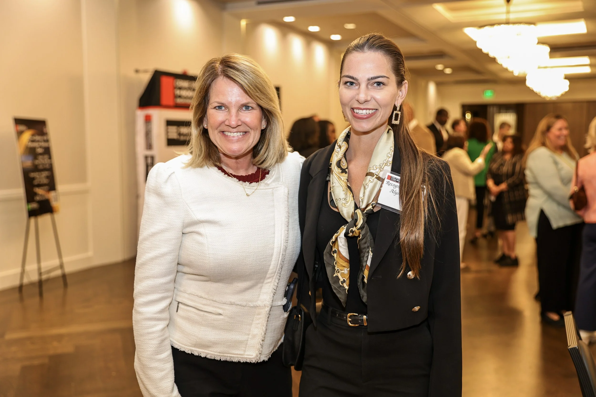 Two women smiling at an indoor networking event, with other attendees in the background, in a well-lit room.