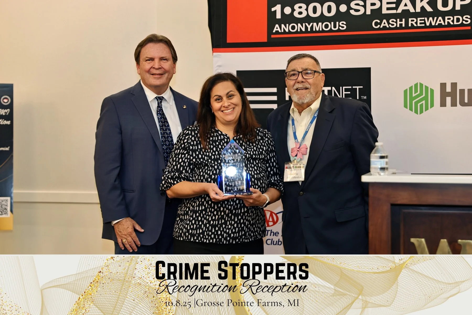 Three people standing together at an event, with a woman in the center holding an award. The man on the left wears a blue suit and tie, the woman in the middle wears a black and white patterned dress, and the man on the right wears a dark suit with a pink bow tie. The backdrop has logos and banners with contact information, and a banner at the bottom reads "Crime Stoppers Recognition Reception, 10.8.25, Grosse Pointe Farms, MI." There is a bottle of hand sanitizer on a table to the right.