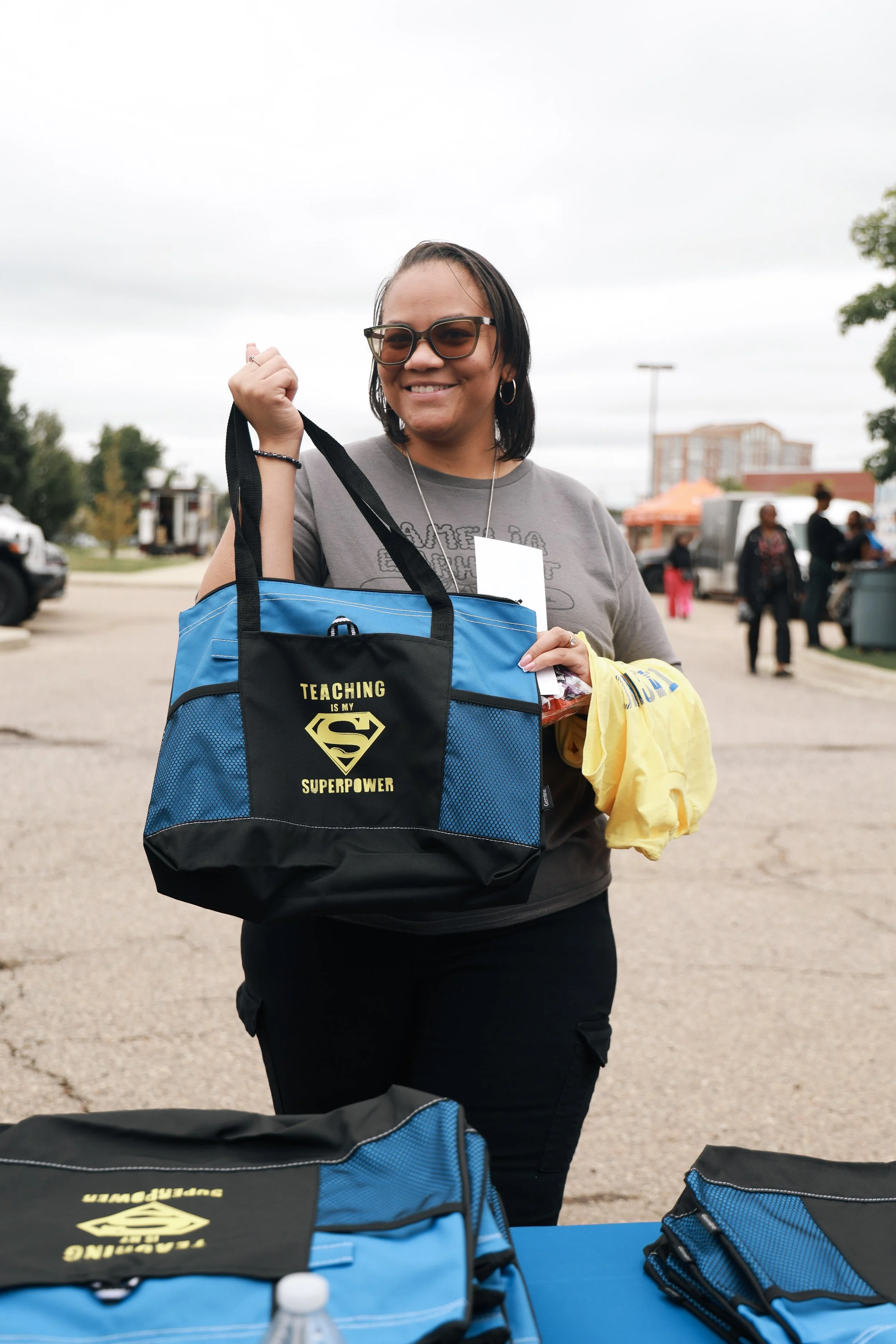 Woman smiling at an outdoor event holding a blue and black tote bag with the phrase "Teaching is my superpower" and a Superman logo, with tables and people in the background.