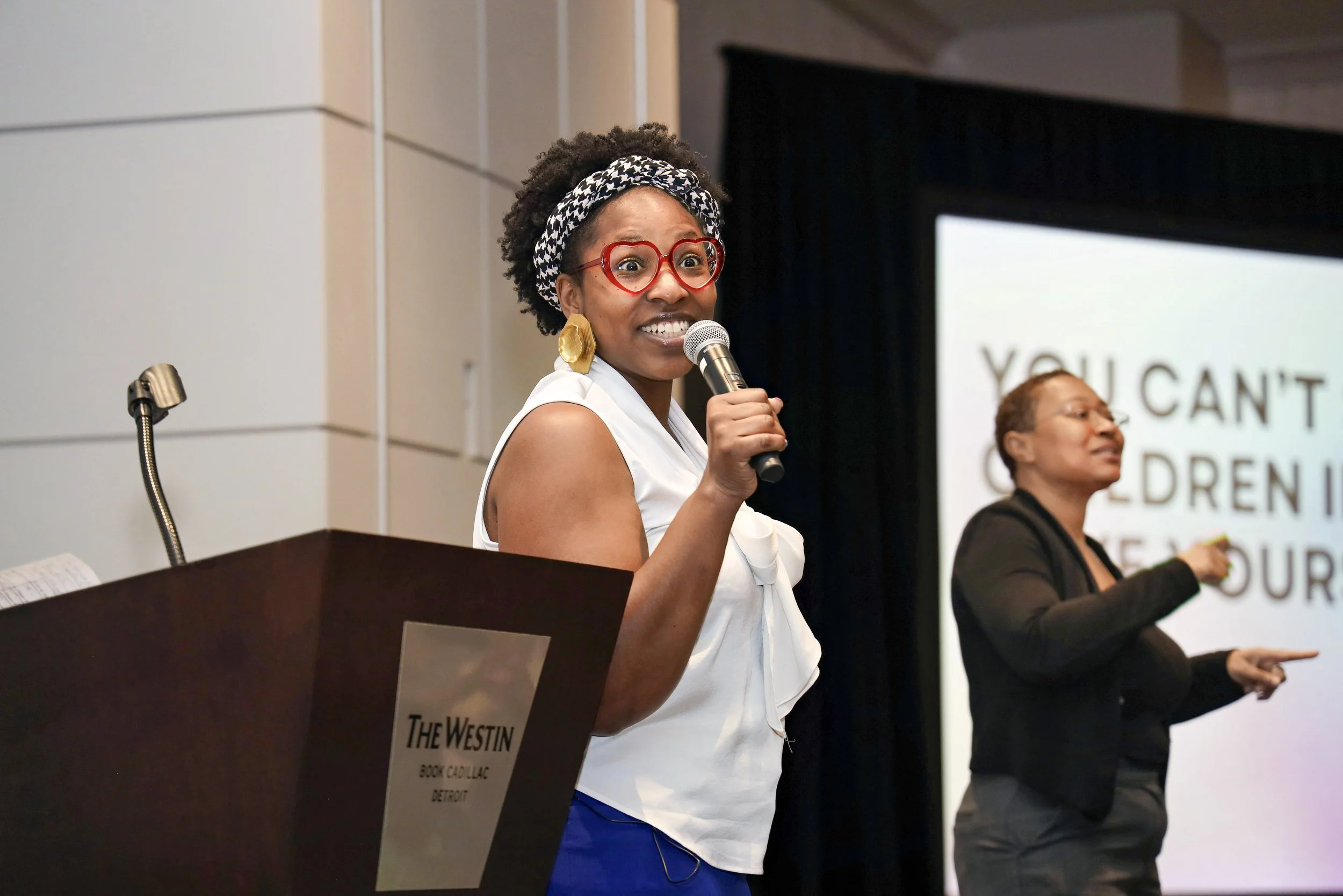 A woman with curly hair, red glasses, and large earrings speaking into a microphone at a presentation in a conference room, with a second woman in the background. The screen behind them displays text about children.