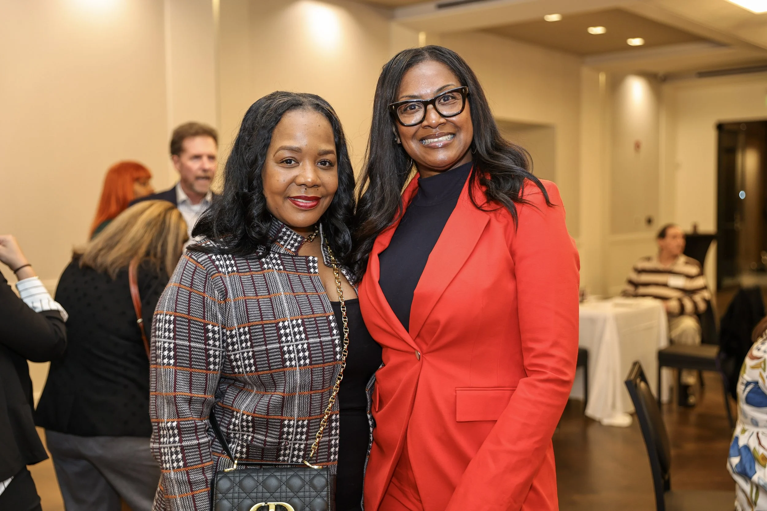 Two women standing together at an indoor event, smiling at the camera. One woman is wearing a patterned blazer and holding a black quilted purse, while the other is dressed in a red blazer with a black top and glasses. In the background, people are m
