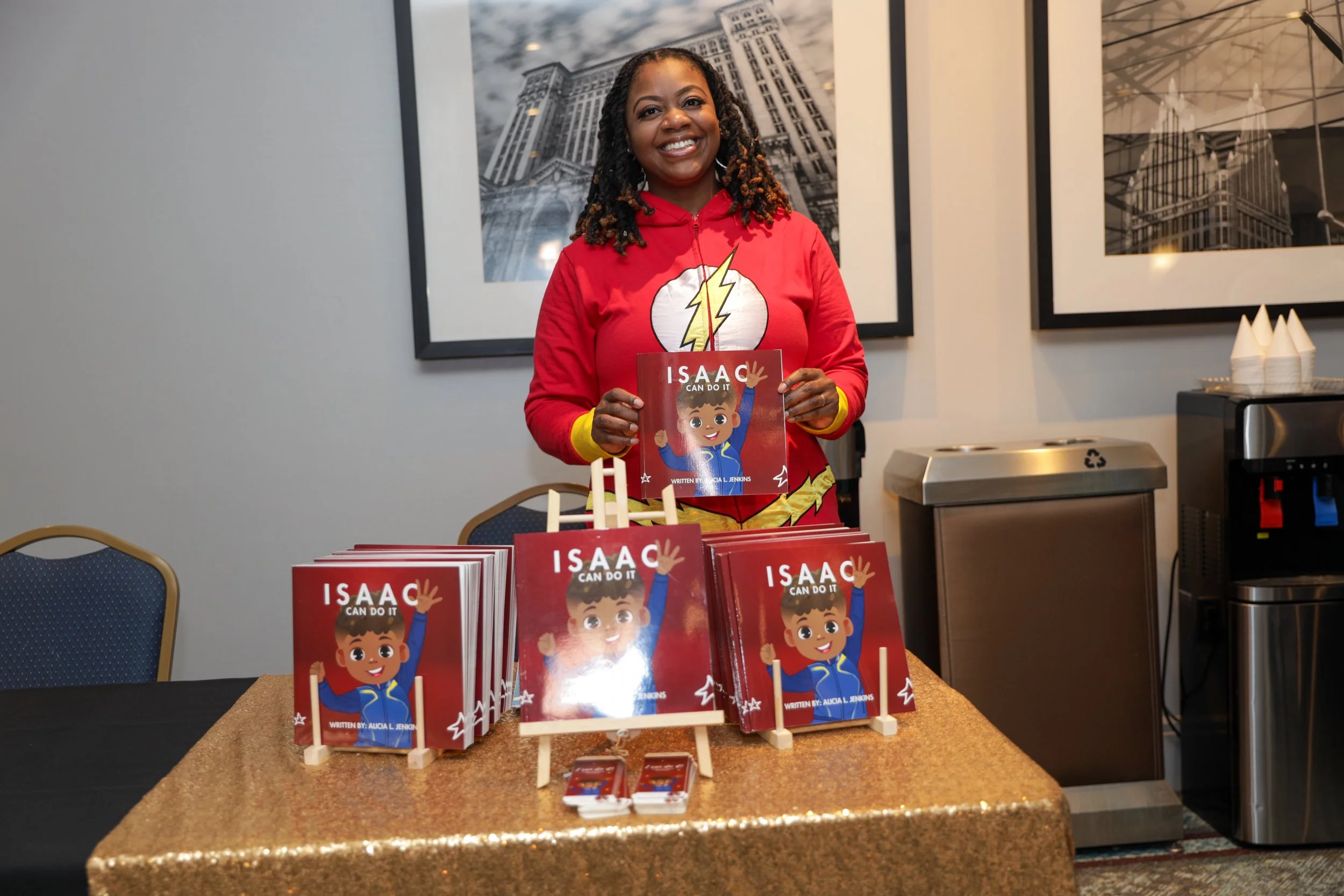 A woman dressed as a superhero in a red costume with a lightning bolt symbol, smiling while holding a children's book titled 'Isaac Can Do It' at a book signing event with copies of the book displayed on a table.