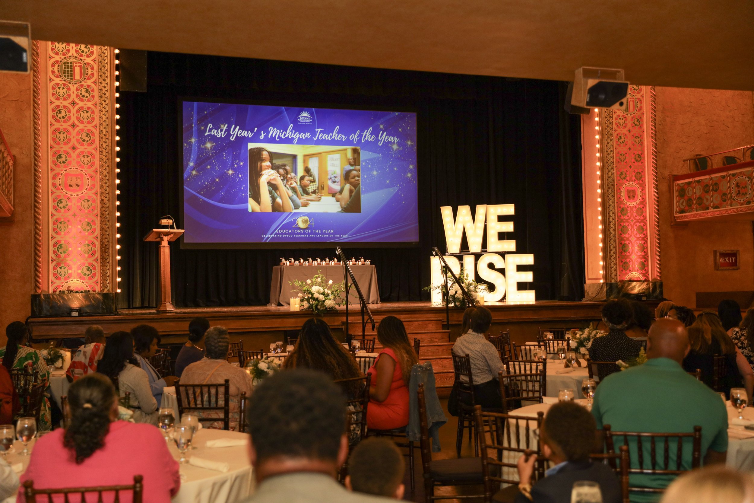 Audience seated at tables watching a stage with a large screen displaying a woman and children, with the text 'Last Year's Michigan Teacher of the Year'. Large illuminated letters spelling 'WE RISE' are on the stage.