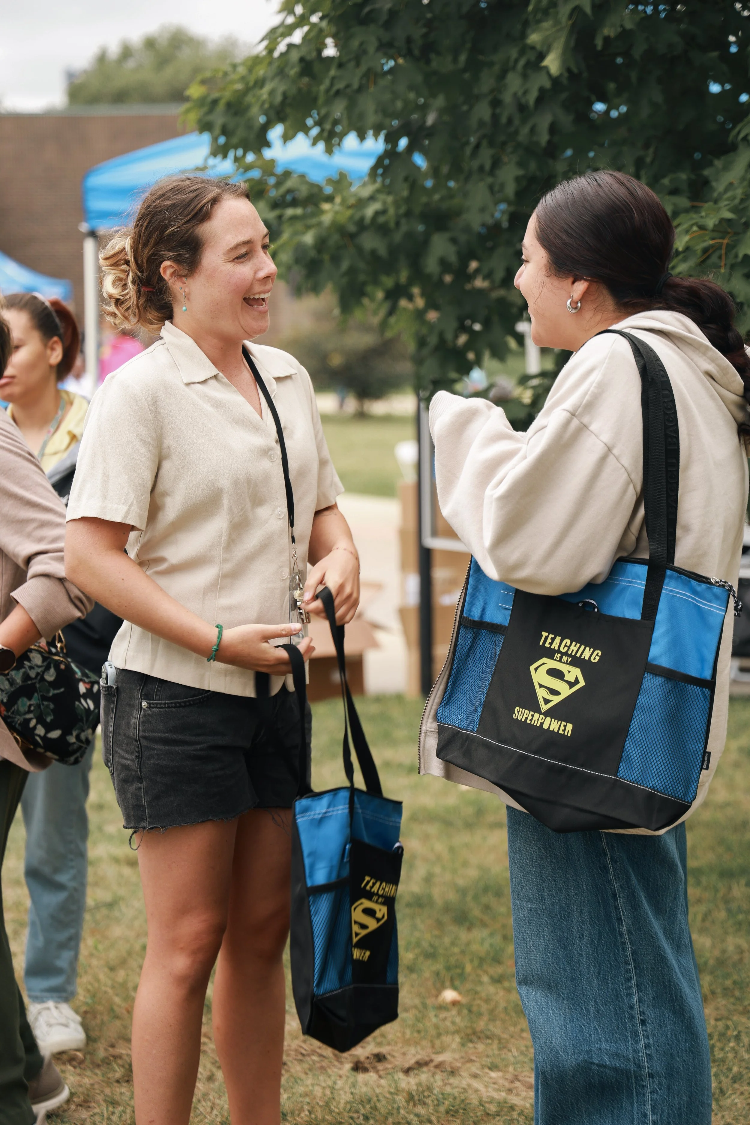 Two women are talking and smiling at an outdoor event, with people and a blue tent in the background. They are carrying tote bags labeled 'Teaching is my Superpower' with a Superman logo.