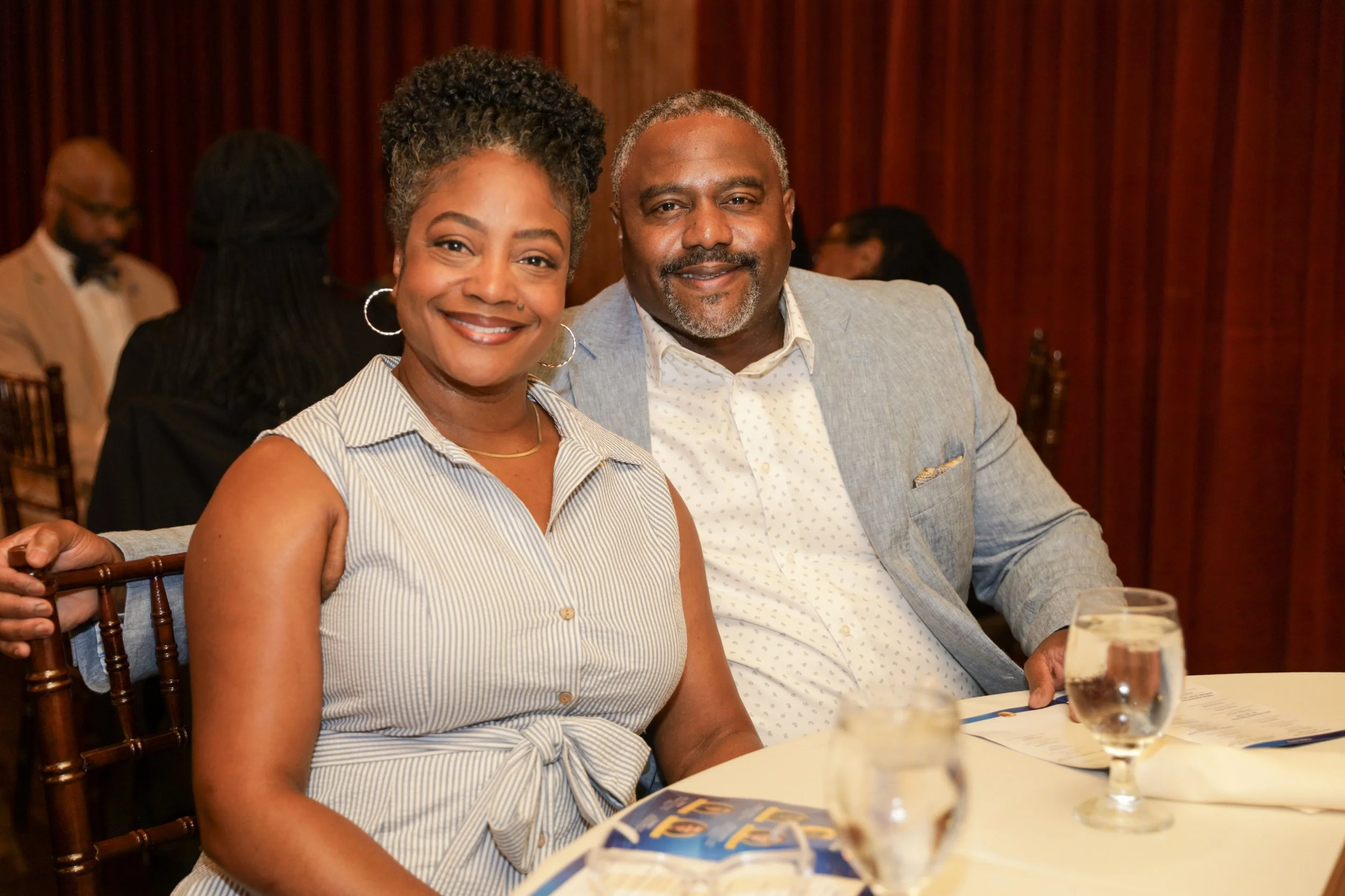 A smiling African American woman in a sleeveless striped top and a middle-aged African American man in a light grey blazer and white shirt sitting at a round table in a formal setting with dark wood paneling in the background.