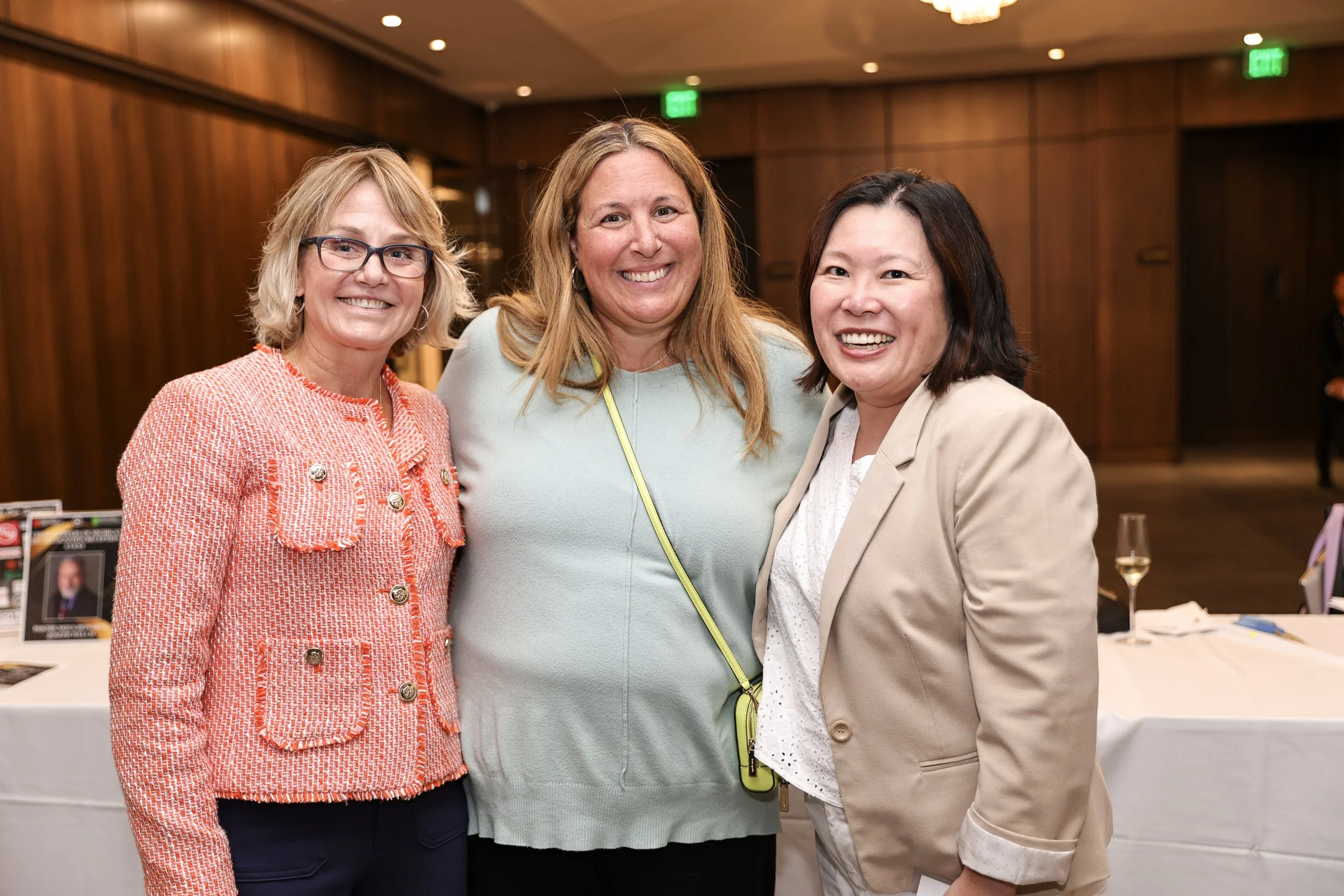 Three women smiling and posing together in an indoor event space with wood-paneled walls, a table with a wine glass and flyers in the background.