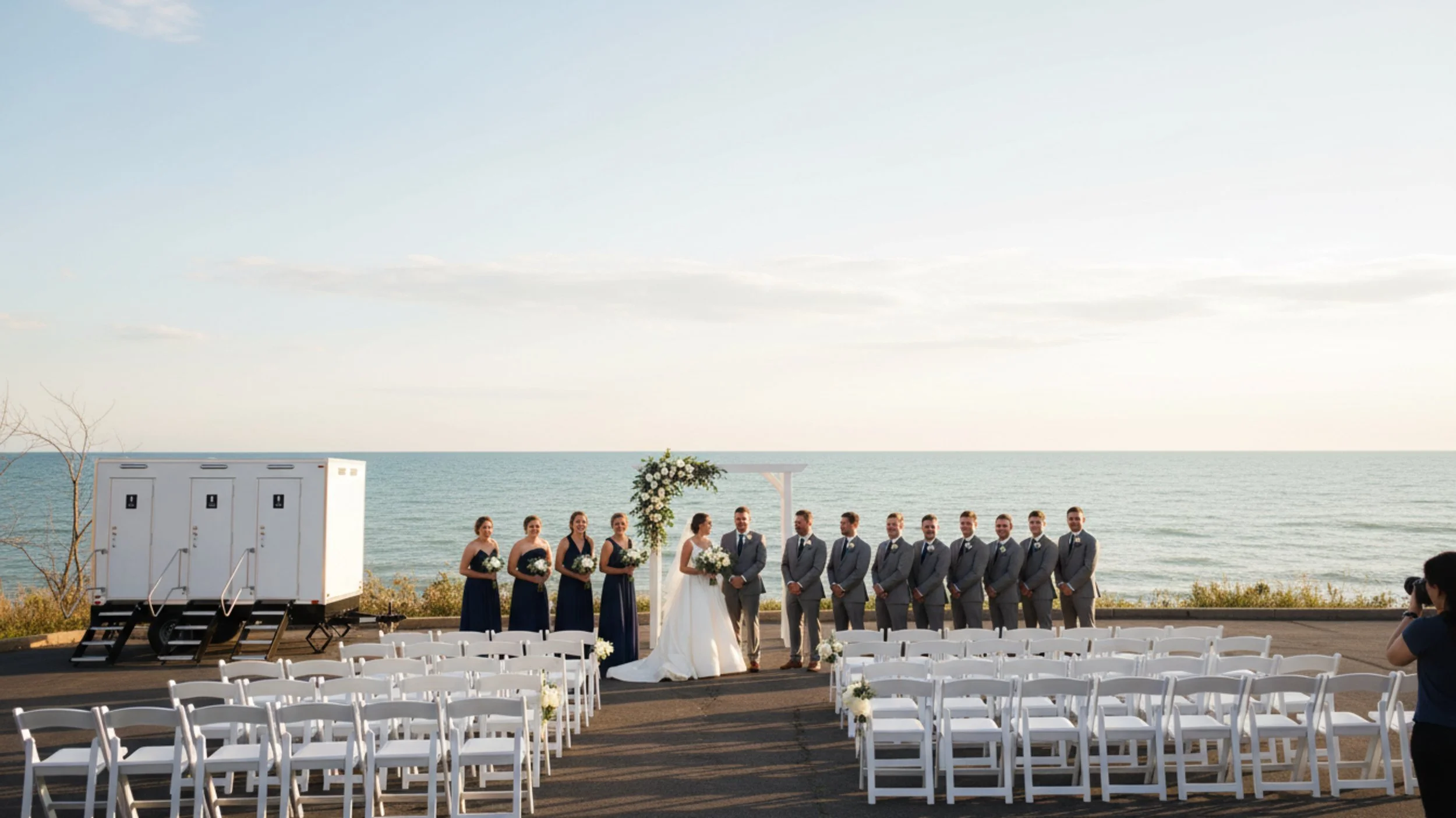 A wedding ceremony takes place outdoors by the ocean, with the bride and groom standing in the center under an arch decorated with white flowers, surrounded by bridesmaids and groomsmen, with white chairs arranged for guests and a photographer taking pictures.