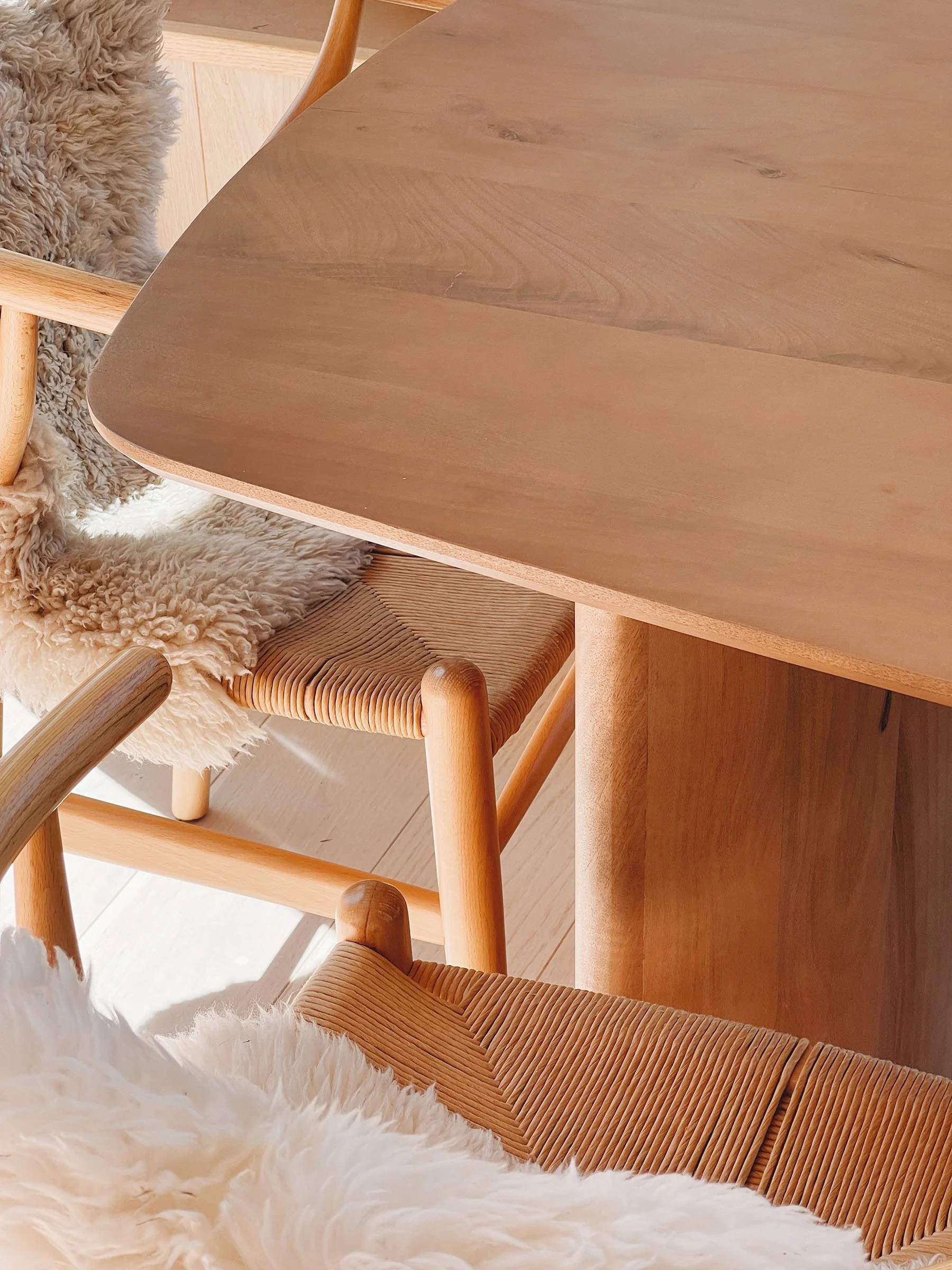 Close-up of a wooden dining table, with chairs around it, one of which has a sheep fur throw on the seat and another with a woven rattan seat.