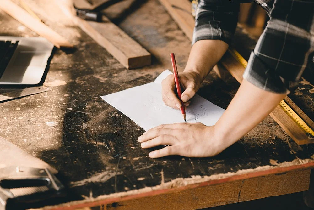 View of man's hands at a woodshop table.  He is drawing a design for an original furniture piece.