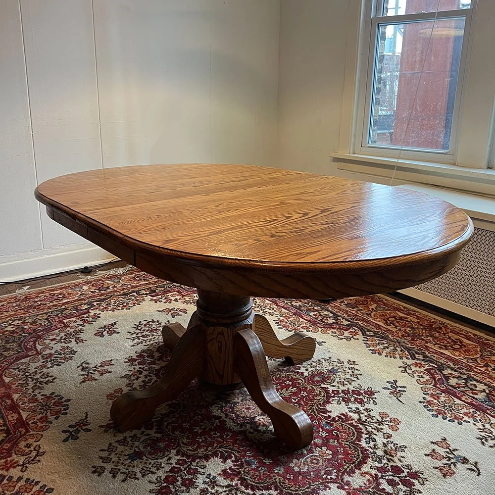 A wooden dining table with a polished surface, placed on a patterned area rug in a room with white walls and a window.