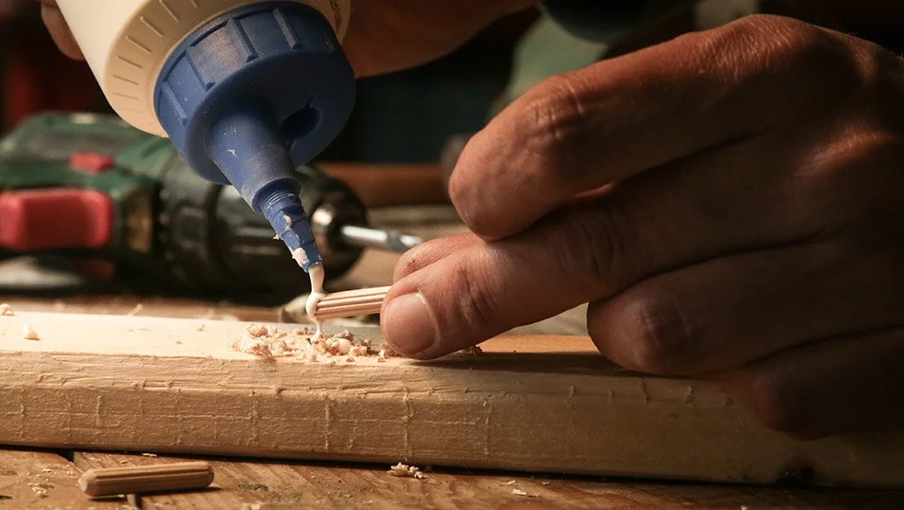 A person applying glue on a small piece of wood in a woodworking project, with a drill in the background.