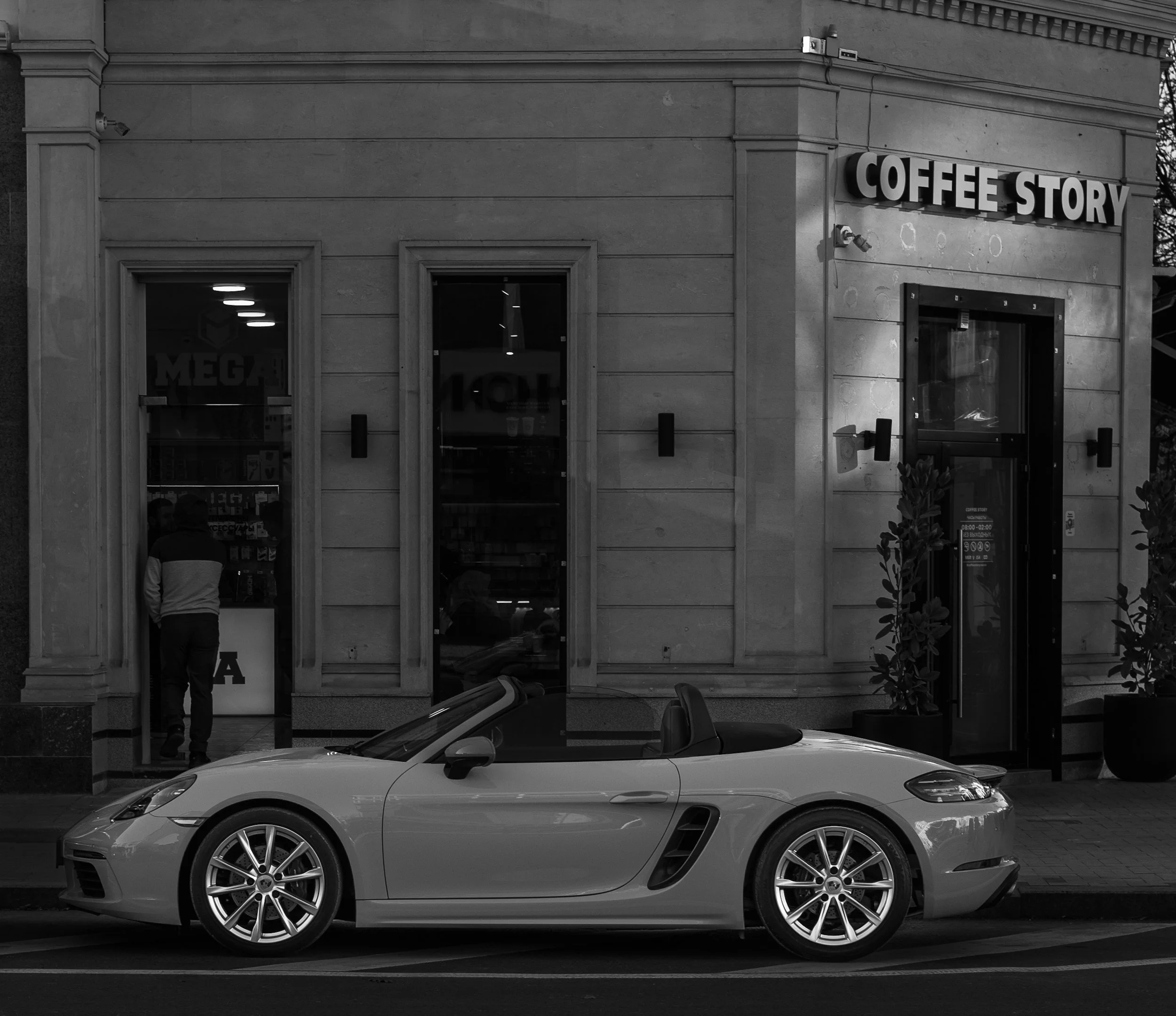 A convertible sports car parked outside a coffee shop with a man inside. The coffee shop has a sign reading "Coffee Story" and appears to be in an urban setting.