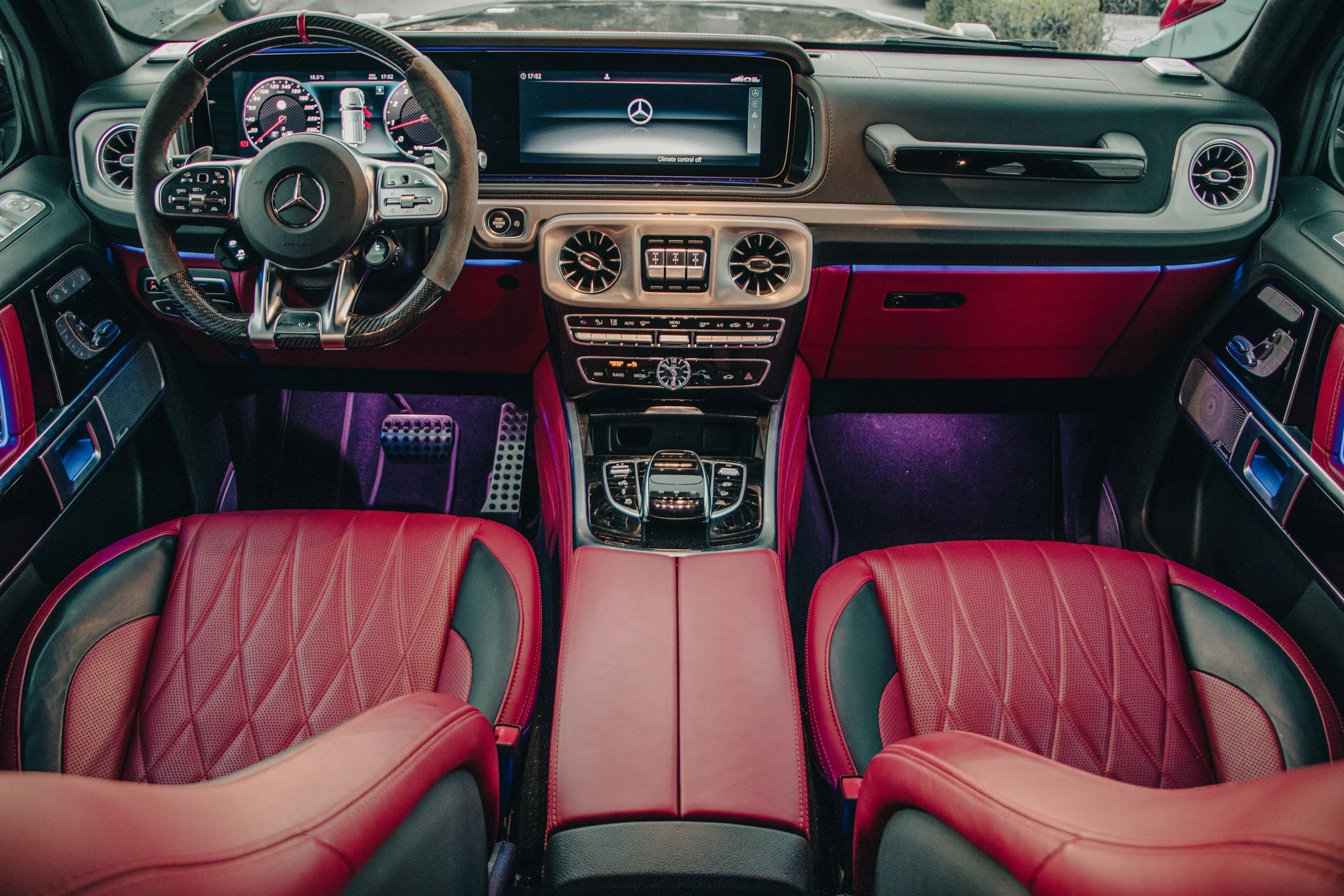 Interior of a luxury Mercedes-Benz vehicle featuring red leather seats, a dashboard with a digital display, and a central console with controls and a clock. The steering wheel has the Mercedes logo and various control buttons.