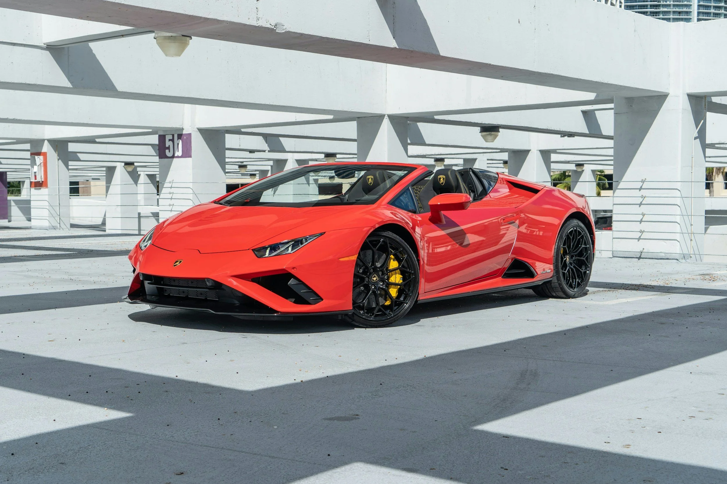 Red Lamborghini sports car parked in a white multi-level parking garage with shadow patterns on the ground.