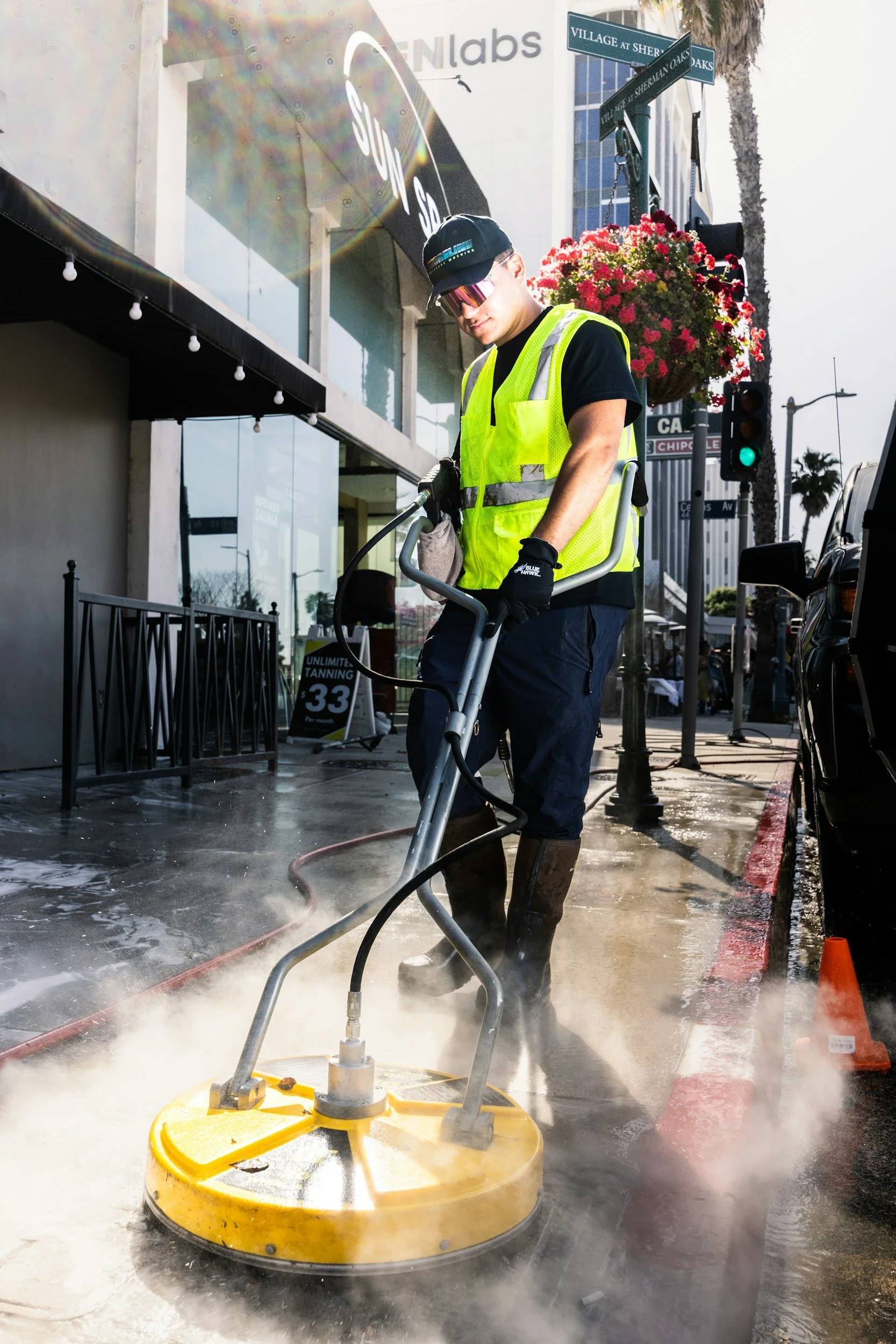 A man wearing a yellow safety vest, black gloves, and boots is operating a steam-cleaning machine on a city sidewalk. He is standing near a street corner with street signs, a traffic light, and a large flower basket hanging from a pole, with a building and palm trees in the background.
