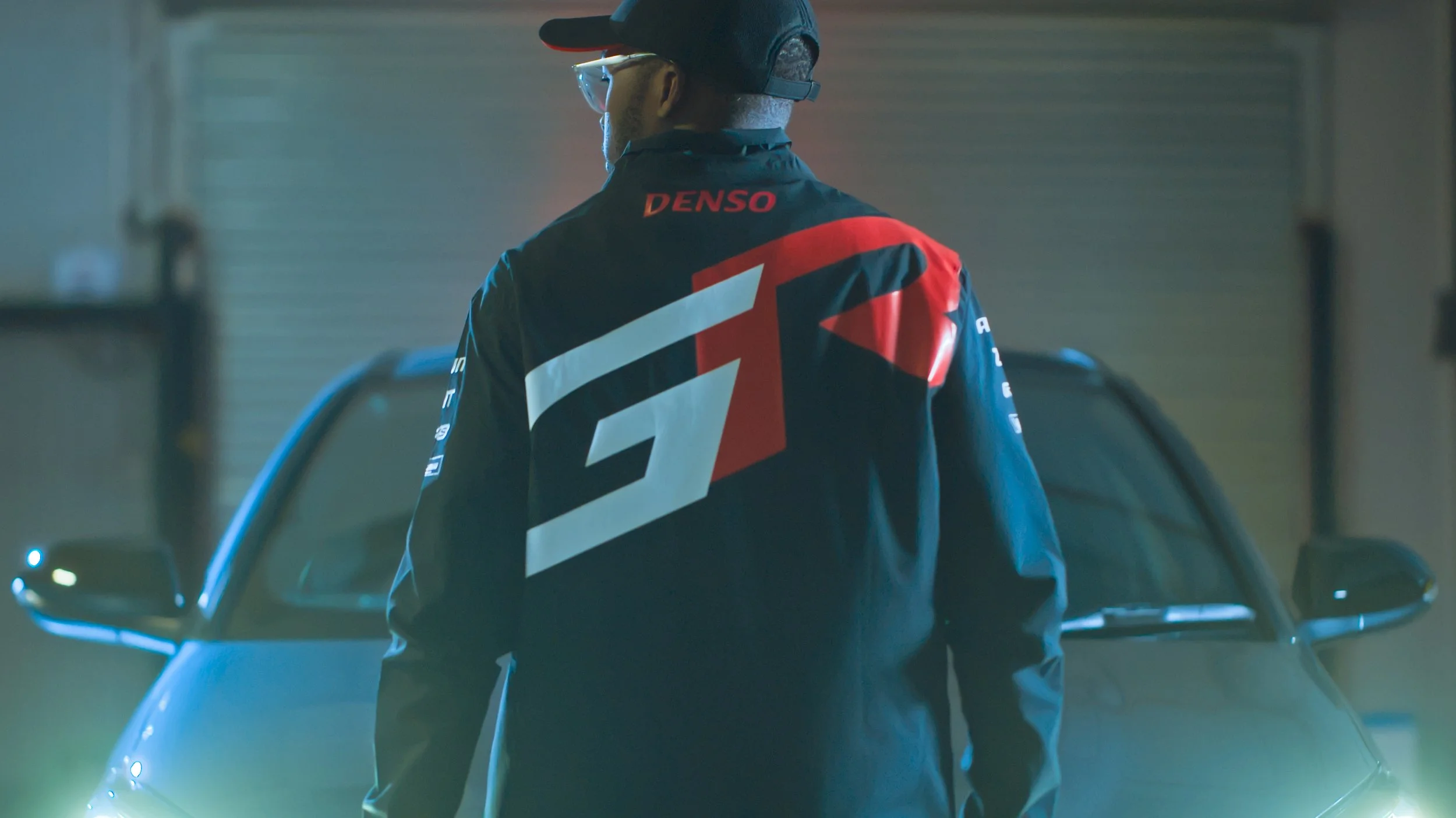 A landscape with a calm lake, mountains in the distance, a few trees, and a modern wooden building in the foreground.