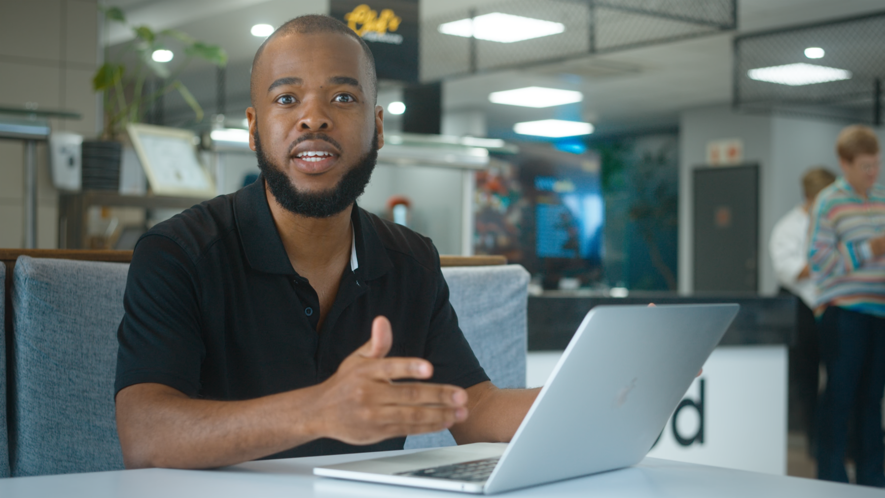 Commercial Cinematography Grab of man sitting at a table with a laptop in a modern indoor setting, gesturing with his right hand and looking at the camera.