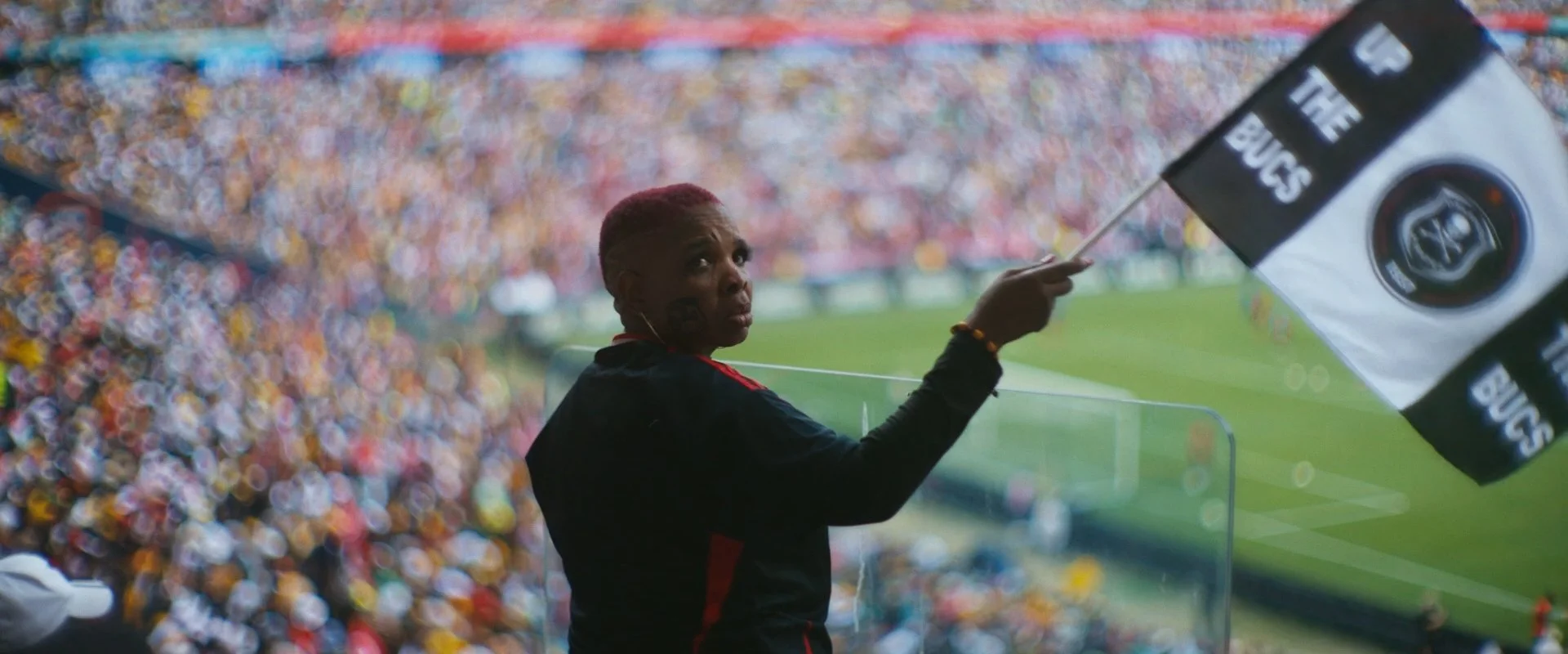 A woman with short hair and earrings at a sports stadium, waving a black and white flag with the Orlando Pirates logo, during the Soweto Derby. Documentary cinematography work. 