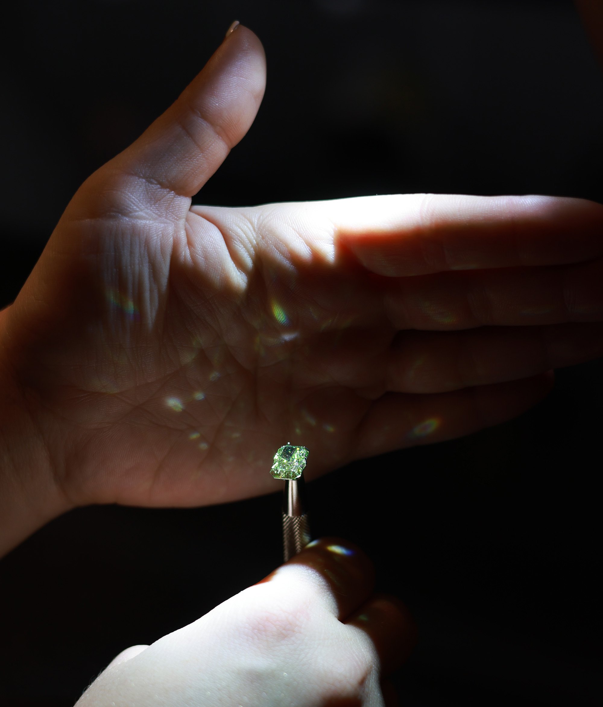 A person holding a ring with a large square-shaped green gemstone, illuminated by a focused light source that creates colorful reflections on their hand and casts a shadow on the background.