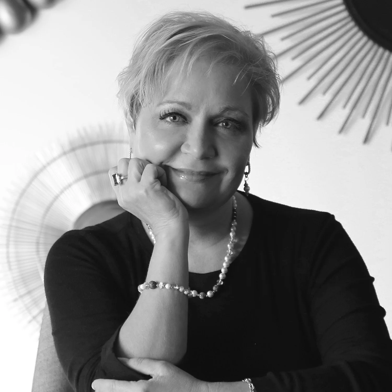 A black and white portrait of a woman with short blonde hair, wearing a black top, pearl necklace, and earrings. She is smiling gently, resting her chin on her hand, with decorative wall art in the background.