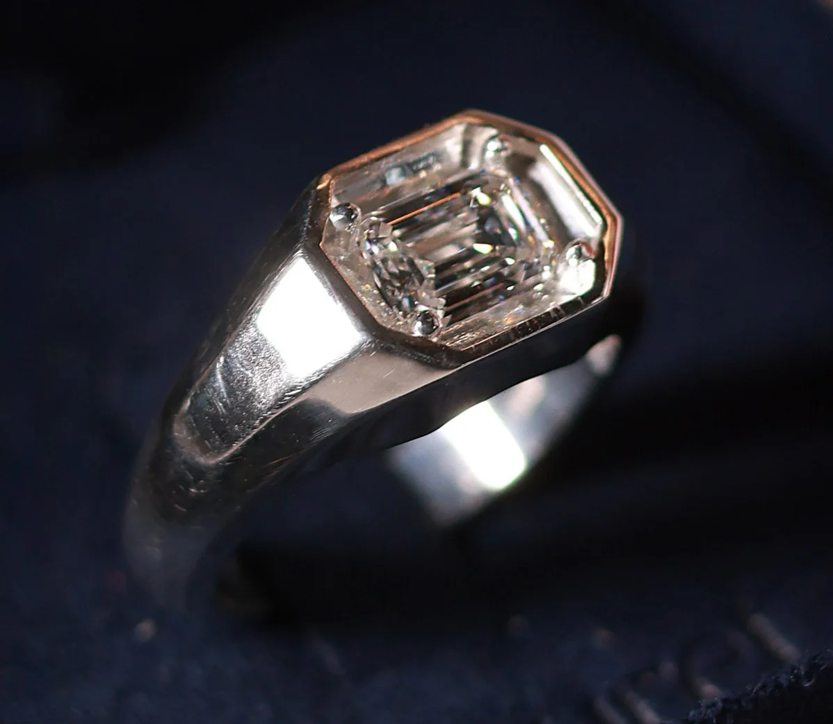 A close-up of a silver ring with a large rectangular-cut diamond set in a bezel setting on a dark fabric background.