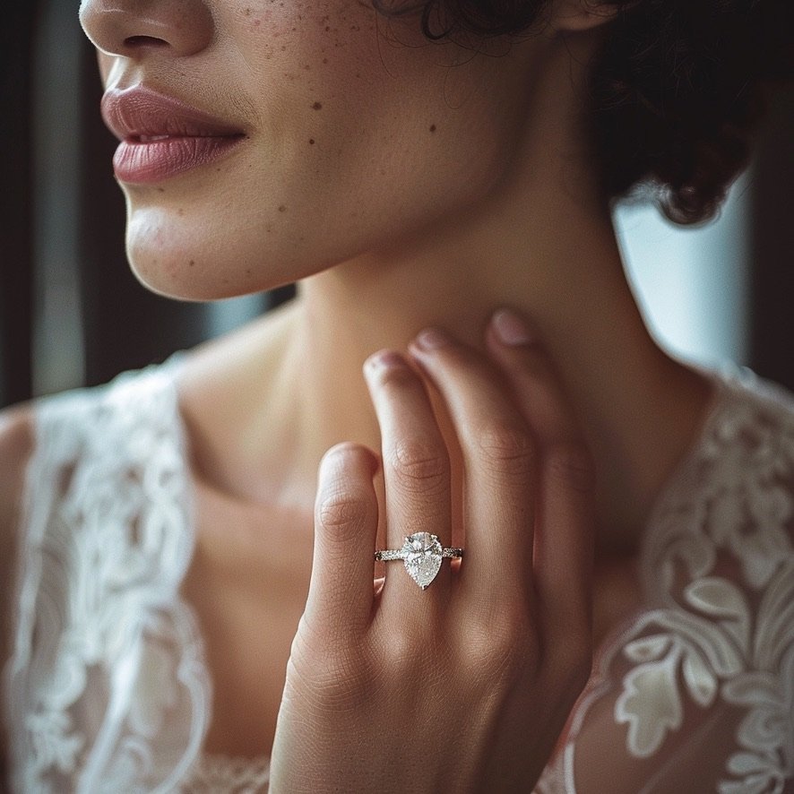 Close-up of a woman's hand with a large heart-shaped diamond engagement ring, touching her neck. She has freckles and is wearing a lace garment.