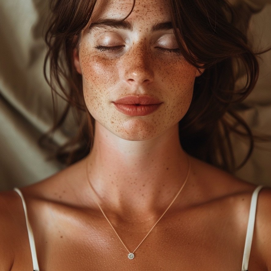 A woman with closed eyes lying down, her face covered in freckles, wearing a necklace with a small pendant and a white tank top.