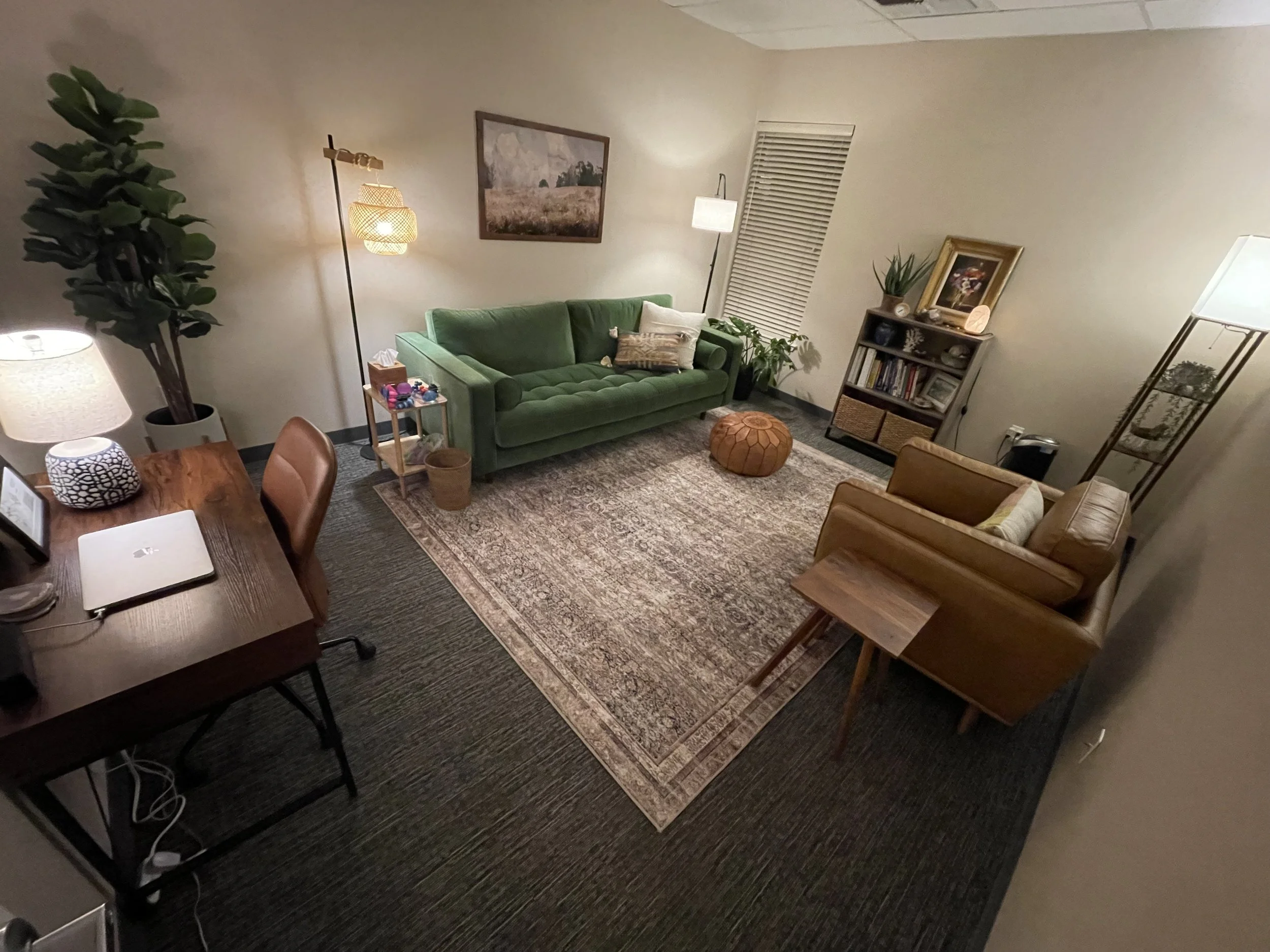 Living room with a green sofa, tan armchair, patterned area rug, and various decorations including plants, lamps, and framed pictures.