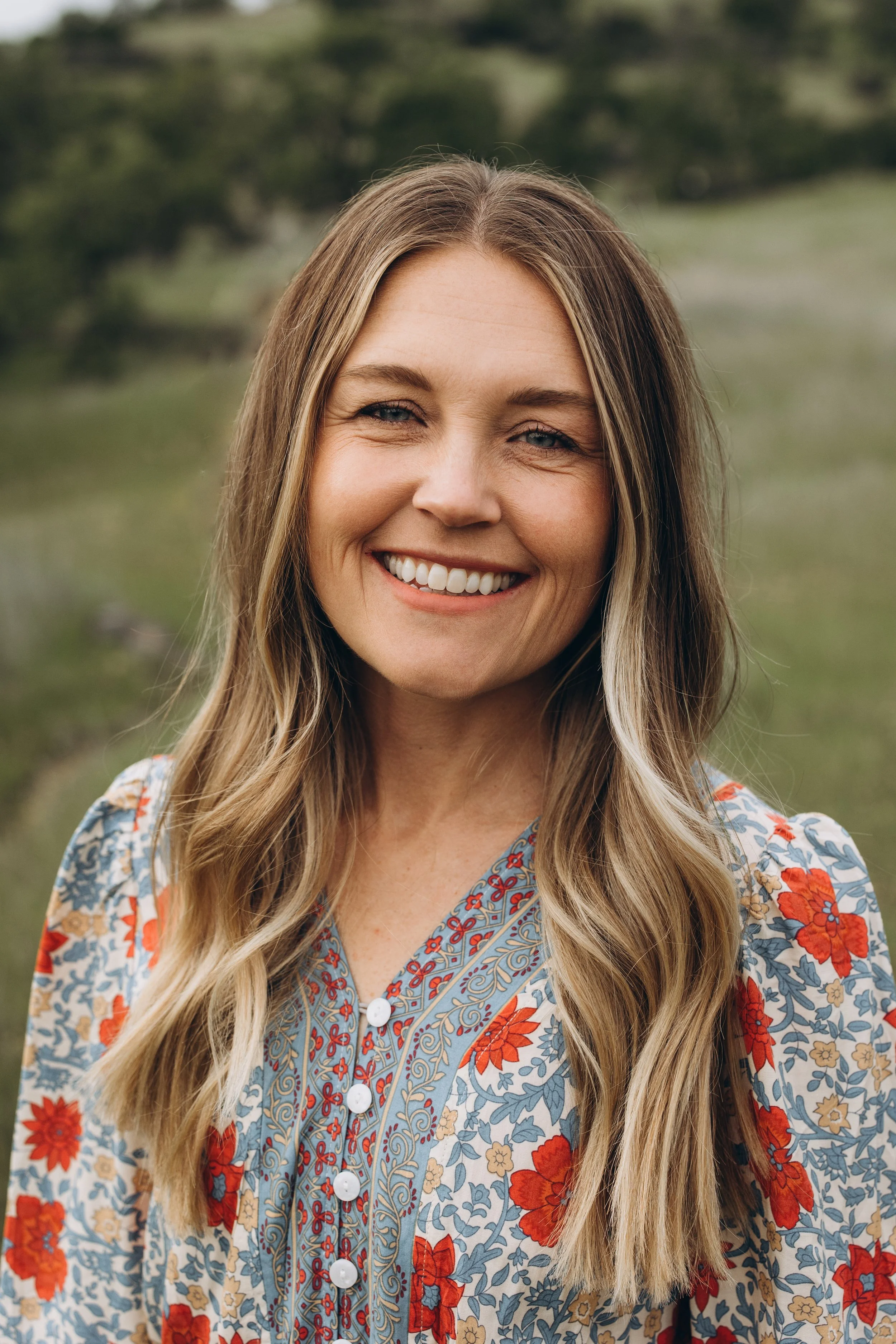 A smiling woman with long blonde hair wearing a floral blouse standing outdoors in a natural setting.