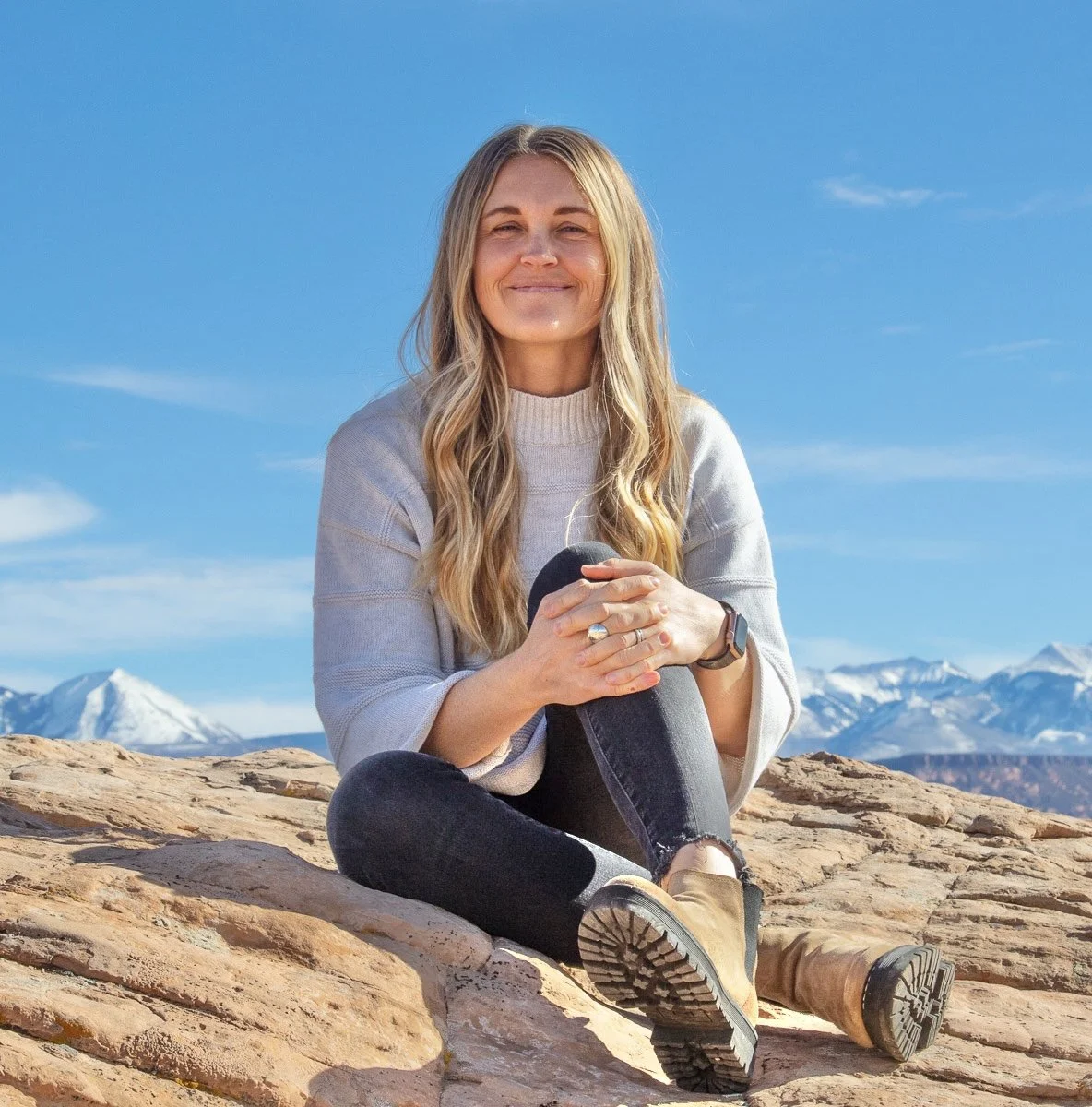 A woman with long blonde hair sitting on a rocky landscape with snow-capped mountains in the background, smiling and holding her hands together.