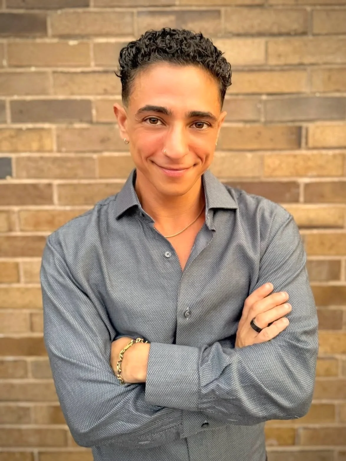 A person with short curly hair and light brown skin, wearing a gray button-up shirt, smiling with arms crossed, standing in front of a brick wall.